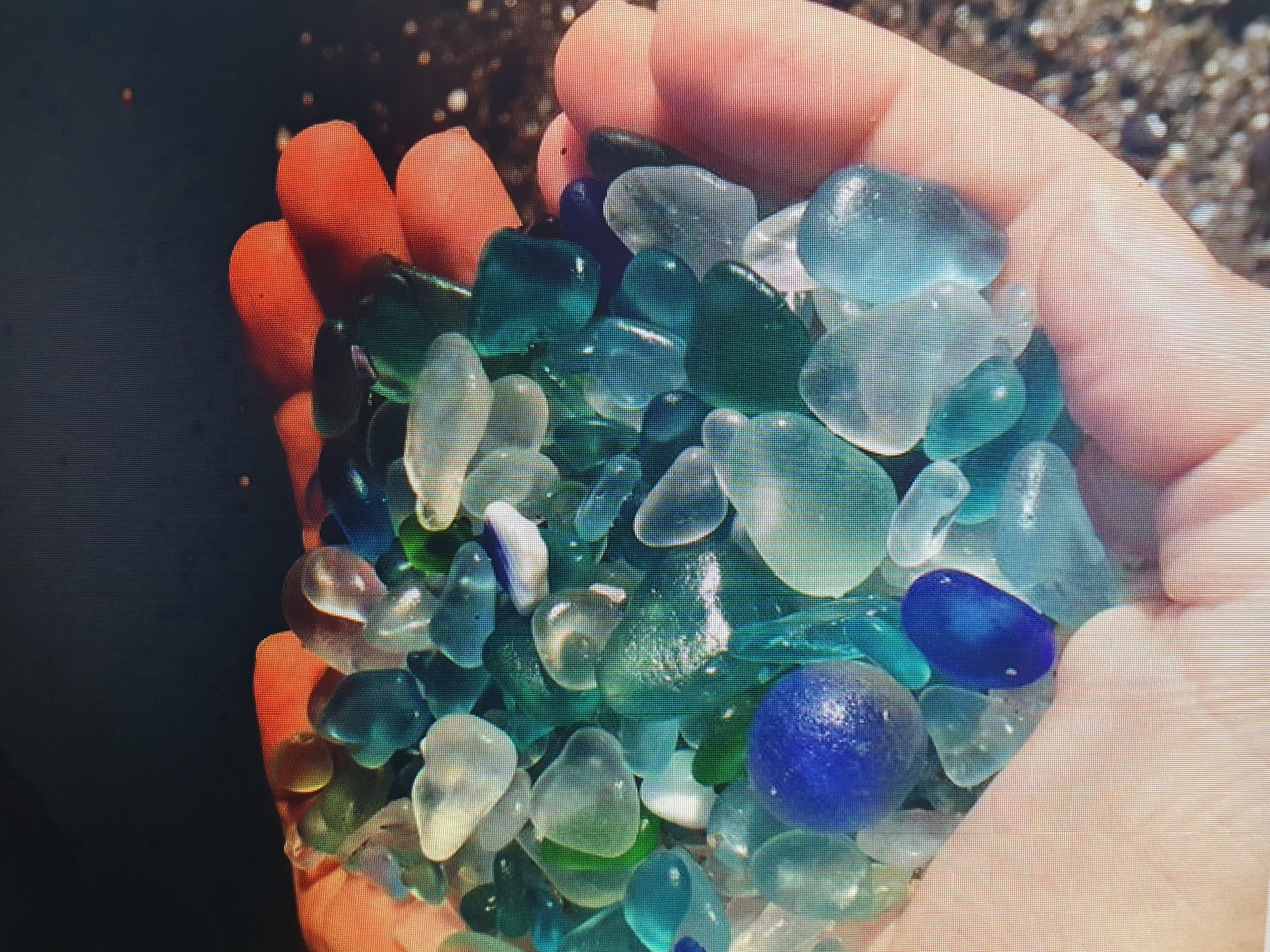 A close-up, overhead shot captures a person's hands cradling a collection of sea glass. The sea glass is varied in size and shape, predominantly in shades of blue, green, and clear, with two prominent deep blue pieces. The hand, a pale skin tone, is gently cupped, holding the glass. The background is dark and out of focus, with some blurred lights that could suggest an outdoor environment or a surface with reflections. No specific location within Tashkent, Uzbekistan, is identifiable from the image.