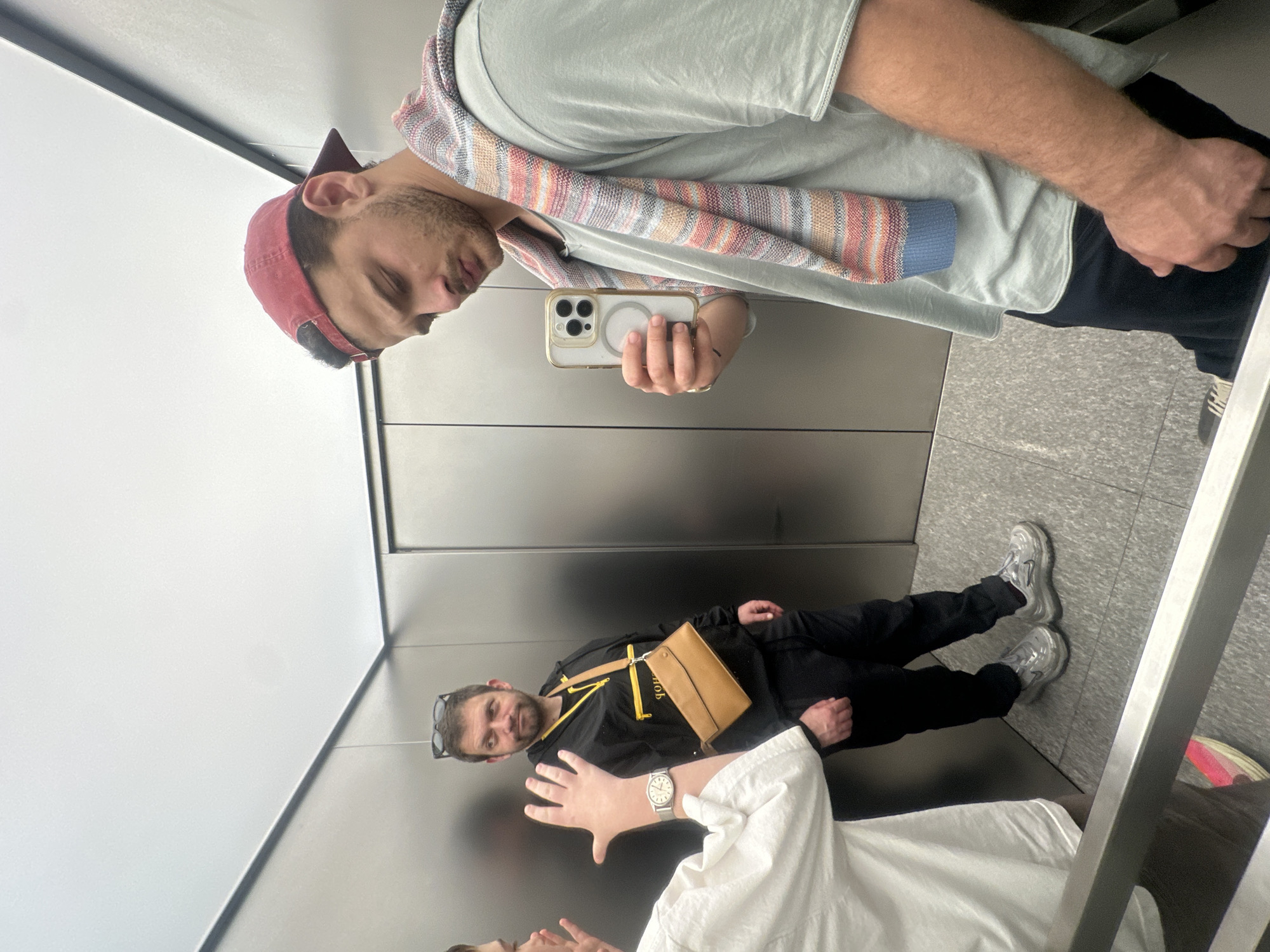 The image shows three individuals inside an elevator. One person is taking a selfie in a mirror. He is wearing a backwards red baseball cap, a light green shirt, and a colorful striped scarf around his neck. He is holding a white iPhone with three camera lenses. Another person is wearing a black hoodie with yellow accents, black pants, and gray sneakers. They have a tan-colored crossbody bag. The third person is wearing a white shirt and part of their face and hand with a silver watch are visible. The interior of the elevator is metallic with gray tiled flooring. The scene appears to be in an elevator within a building in Milan, Italy.