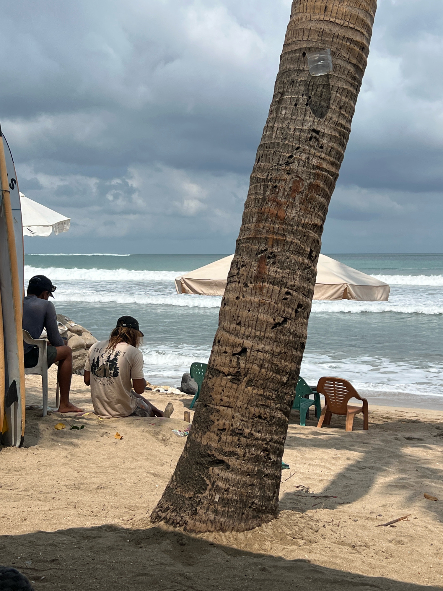 Two individuals are seated on a sandy beach in Kuta, Indonesia, facing the ocean. The person on the left is sitting on a white plastic chair, wearing a dark grey long-sleeved shirt and dark shorts, with a black baseball cap. They are looking towards the ocean. The person on the right is sitting on the sand, wearing a light-colored t-shirt with a graphic design and patterned shorts, also with a black baseball cap. They appear to be looking down. A surfboard stands upright to the left of the frame. In the background, waves crash on the shore under a cloudy sky. Two beach umbrellas are visible further back, one white and one tan. A large palm tree trunk dominates the foreground on the right side of the image. Two empty plastic chairs, one green and one brown, are placed on the sand closer to the water. The scene is set on a beach with light brown sand, scattered with some small debris.