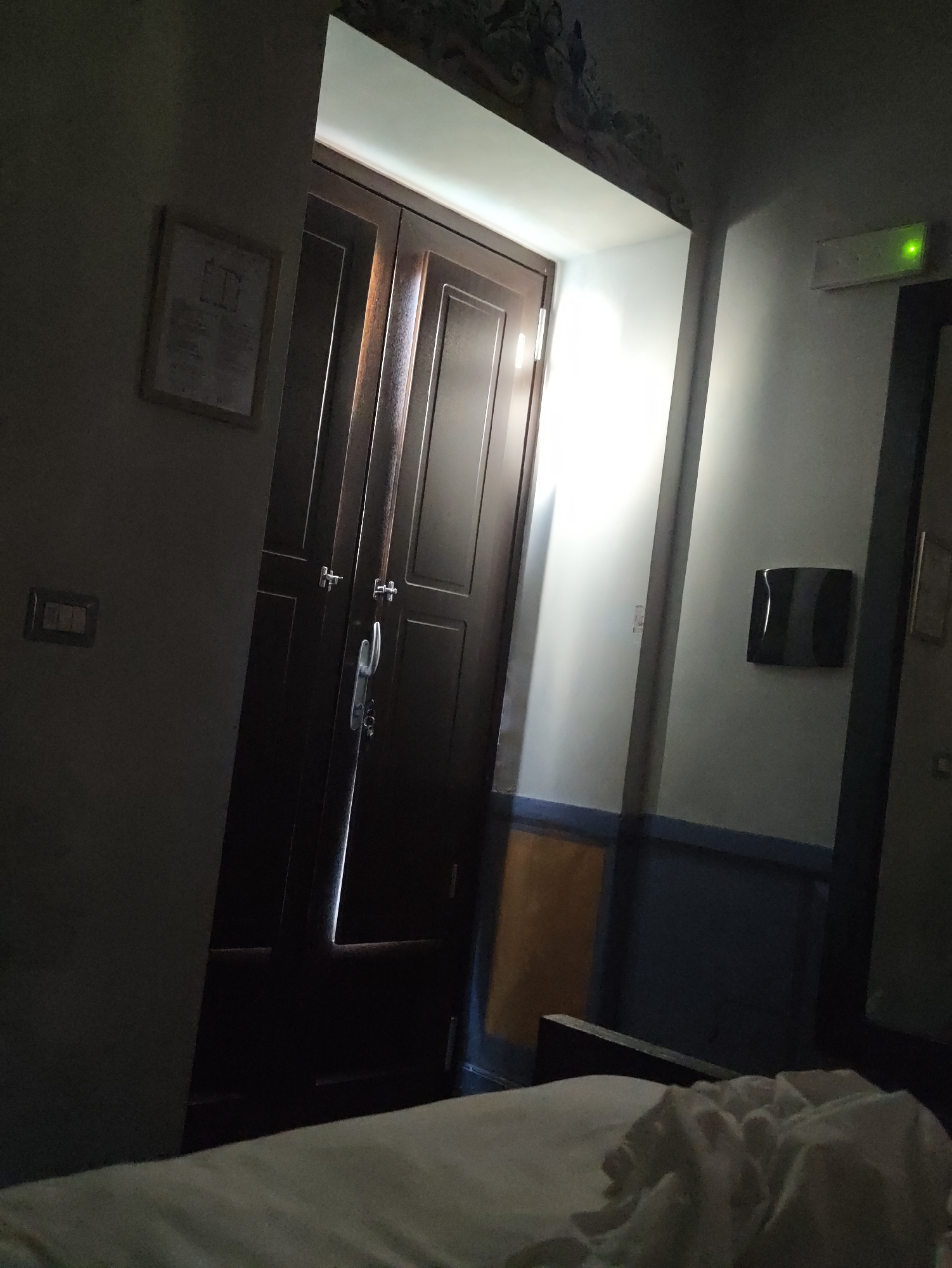 The image depicts an interior scene, likely a hotel room, in Pozzuoli, Italy. Dominating the left side of the frame is a large, dark wooden double door with ornate handles. A sliver of bright light emanates from the crack between the doors, suggesting an open space beyond. To the left of the door, a framed document is affixed to a pale gray wall. Below the document, a light switch plate is visible.
The right side of the image shows a white wall with a blue and orange horizontal stripe pattern near the bottom. A black, rectangular object, possibly a ventilation grille or a decorative panel, is mounted on the wall. Further to the right, a green emergency exit light is illuminated. The foreground of the image is occupied by a rumpled white sheet, suggesting an unmade bed. The lighting is dim, with significant shadows cast across the scene. The ceiling features a decorative mural, partially obscured by darkness.
