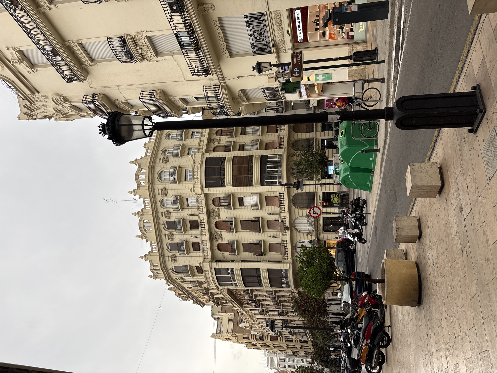 This image captures a street scene in Valencia, Spain, likely in a commercial district given the storefronts. The architecture features buildings with ornate facades, cream-colored paint, and balconies with black wrought-iron railings. A tall, black streetlamp stands prominently in the foreground, to the left of center.
On the sidewalk, there are concrete planters, one with a small tree. Several concrete blocks are also present. A row of parked motorcycles and scooters lines the street on the left. Across the street, there are green recycling bins and a bicycle parked next to them.
A street sign indicates "mercado c. Colón", suggesting proximity to the Central Market of Colon. Storefronts visible include "Blue Banana" and "Le Creuset", suggesting a shopping area. Two individuals are visible near the storefronts. The sky is overcast, contributing to a muted lighting condition. The street is paved with asphalt, and there are white lane markings. The overall impression is a bustling urban environment with a blend of historic architecture and modern amenities.