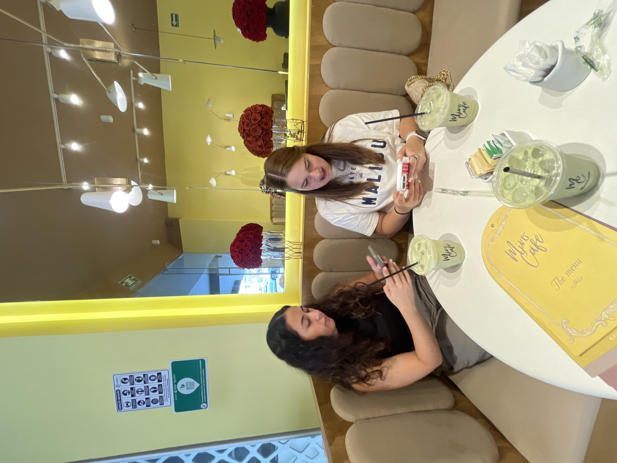 Two young women are seated around a white, round table in what appears to be a cafe interior. The woman on the right has long, wavy, dark hair and is wearing a black top. She is looking at her smartphone. The other woman, on the left, has medium-length brown hair and is wearing a white T-shirt with the word "MALIBU" printed on it. She's holding what appears to be a white mobile phone.
On the table are several green-colored drinks in clear cups, labeled "Mino's Cafe," a small container of sugar packets, a white container of tissues, and a large, yellow menu.
The background is yellow with decorative lighting fixtures and floral arrangements. Safety signs are affixed to the wall.
Based on the menu on the table, the location is Mino's Cafe. While it is not visible in the image, Mino's Cafe is located in the city of Puebla, Mexico.