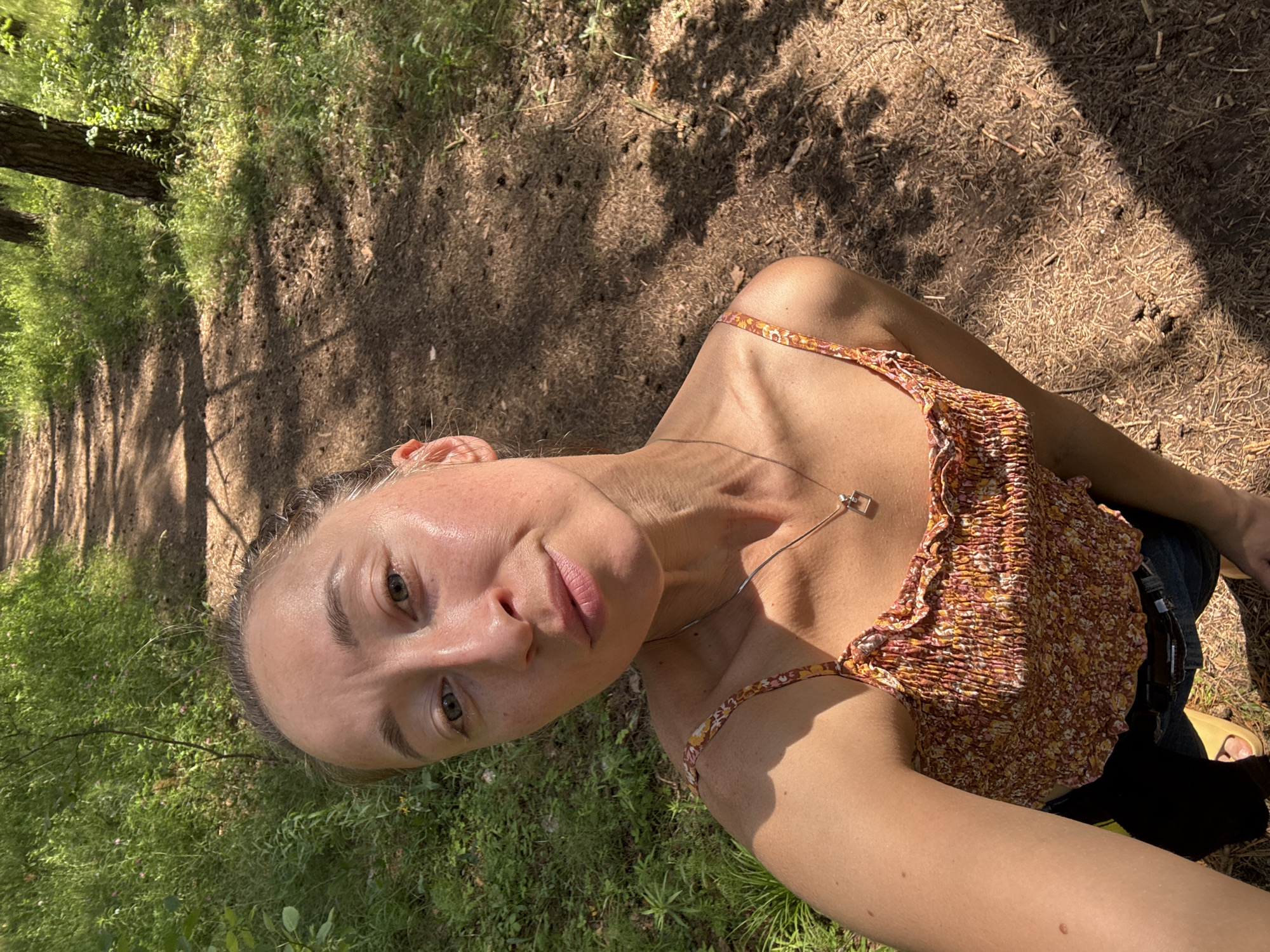 In the city of Perm, Russia, a woman is captured in a selfie taken outdoors on a path in a wooded area. She is wearing a floral-print crop top and denim jeans, with a silver necklace featuring a square pendant. The setting appears to be a forest or park, with a dirt path winding through lush green vegetation and trees. Sunlight filters through the trees, casting dappled shadows on the ground, indicating it is daytime. The woman's expression is neutral as she looks directly into the camera.