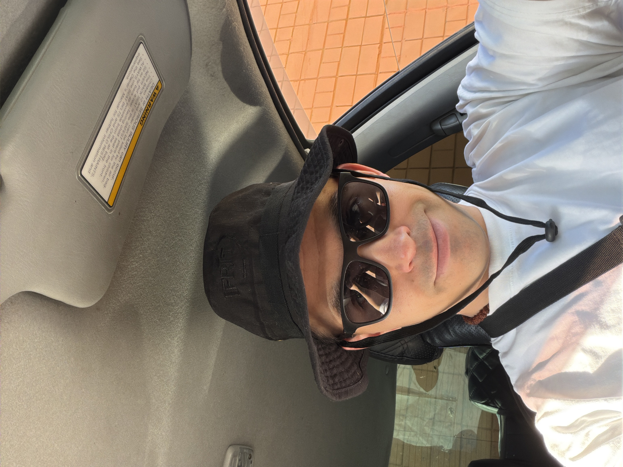 The image shows a man in the driver's seat of a car. He is wearing a white shirt, sunglasses, and a black hat. A warning sticker is visible on the sun visor. The location cannot be determined based on the visual information provided.
