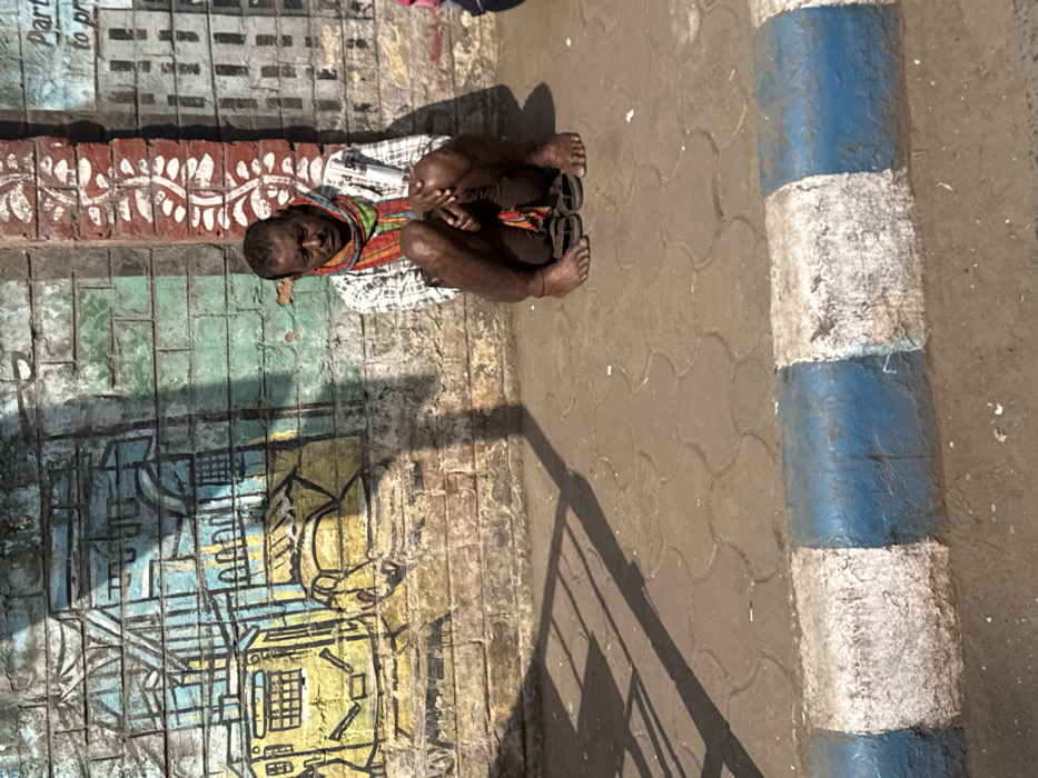 A person sits on the ground next to a wall adorned with colorful graffiti. The individual has dark skin and is wearing sandals, a checkered shirt, and a multi-colored scarf. Part of a pole painted with alternating blue and white stripes is visible on the sidewalk. A shadow is cast diagonally across the ground. The painting features an abstract design in blue, yellow, green, and red.