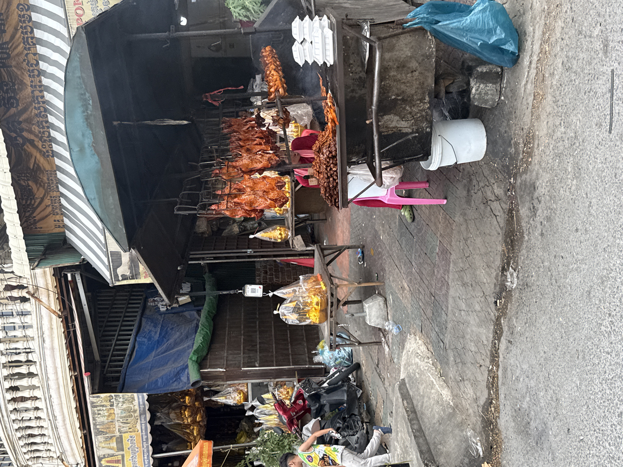 The image depicts an outdoor street food stall in Khan Chamkar Mon, Cambodia, likely during daylight hours. The stall features a variety of food items, primarily roasted ducks and other meats, displayed on racks and a cart. A vendor, wearing a red garment, is partially visible behind the cart. In the foreground, a young boy in a yellow and blue athletic jersey walks on a concrete walkway. Scooters are parked to the left of the stall. The environment appears to be an urban street with weathered pavement and various merchandise displayed at adjacent shops. There are plastic bags, containers, and a white bucket visible around the stall.
