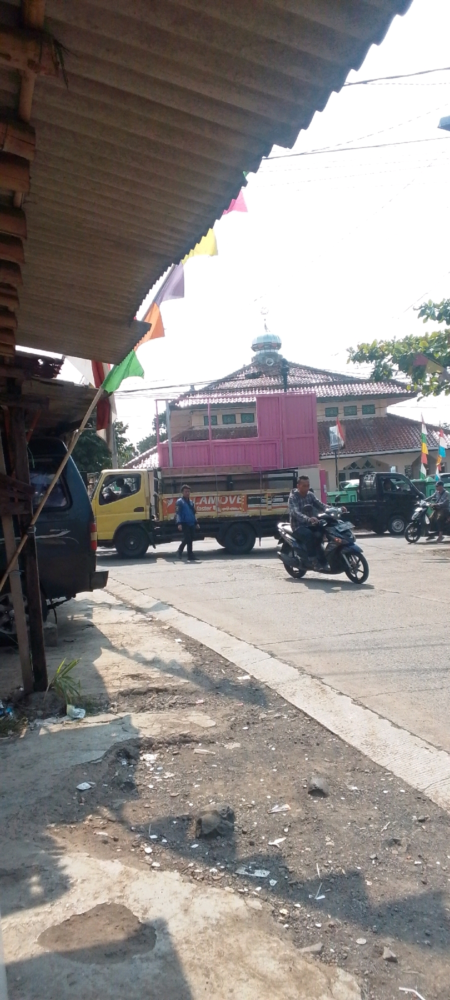 The image depicts a street scene in Cilaku, Indonesia. A pink building with a domed roof, likely a mosque, is visible in the background under a bright sky. In the foreground, a yellow truck with a black flatbed is parked on the road. A person in a blue jacket is standing near the truck, and another person is riding a motorcycle in front of the truck. To the left, a dark-colored van is partially visible. The road surface is worn and appears to have some debris. Colorful flags are strung across the top of the image, suggesting a festive occasion or event. The overall environment appears to be an urban or semi-urban setting.