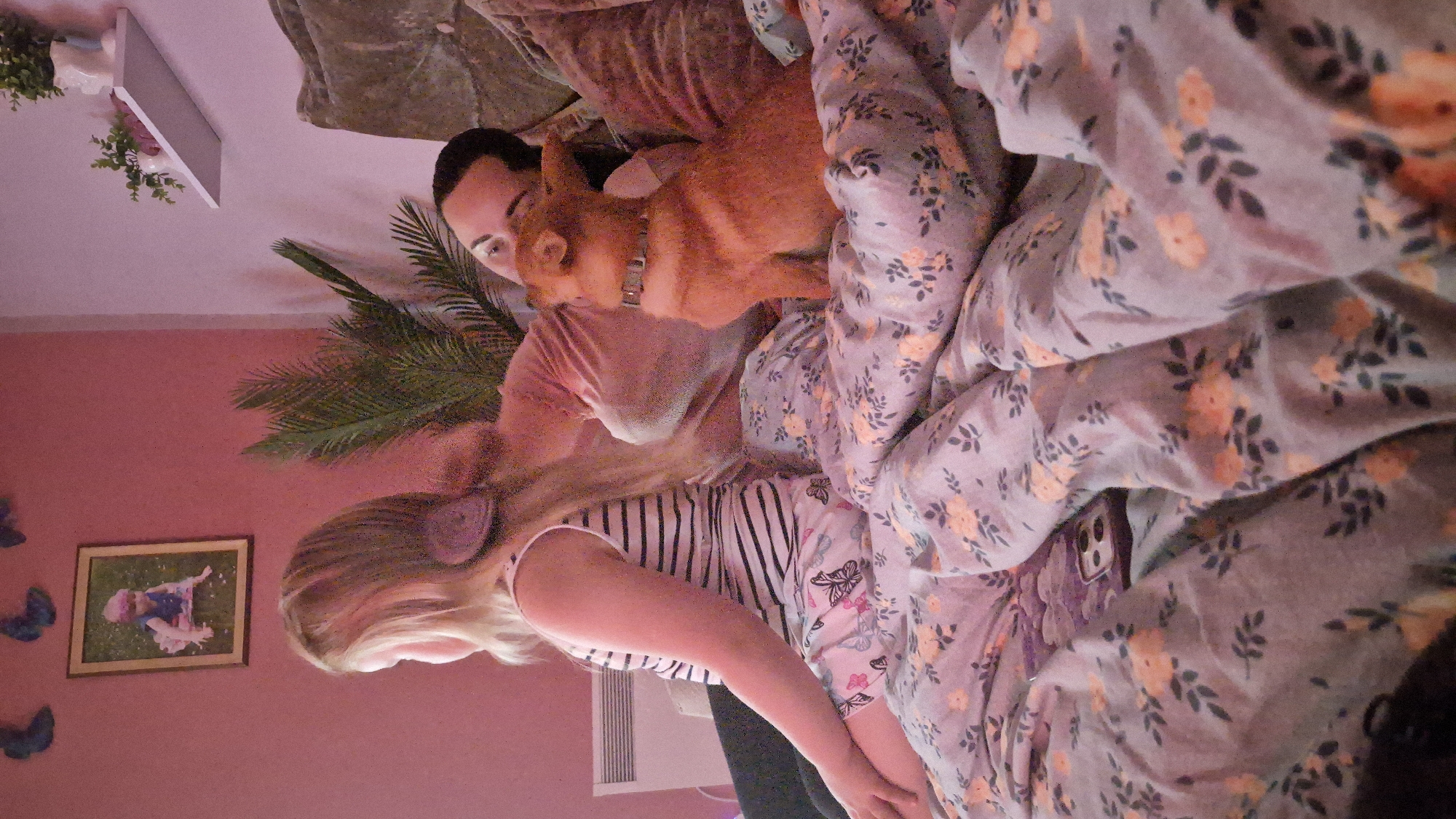 The image depicts a candid, indoor scene with three subjects: a woman, a young girl, and a dog. They are all lying down, seemingly on a bed, under a duvet with a floral pattern in shades of grey and pink. The overall lighting suggests it might be daytime, but the indoor setting with its warm, soft illumination creates a cozy and relaxed atmosphere. The woman, with dark hair, is partially visible, her head resting near the dog. The dog, which appears to be a small to medium-sized breed with reddish-brown fur, is lying on top of the woman, looking towards the side. The young girl, with blonde hair and wearing headphones, is positioned in front of the woman and the dog, facing left. She is wearing a striped shirt.
In the background, on a pink wall, there is a framed picture of a young child. A decorative palm frond is positioned between the woman and the girl. To the left of the framed picture, there are several blue butterfly decorations on the wall. A white shelf with some plants and decorative items is visible in the upper left corner of the image. The scene appears to be a bedroom or a living room, given the presence of a bed-like surface and home decor. The emotion conveyed is one of comfort and togetherness. There is no visible text in the image.