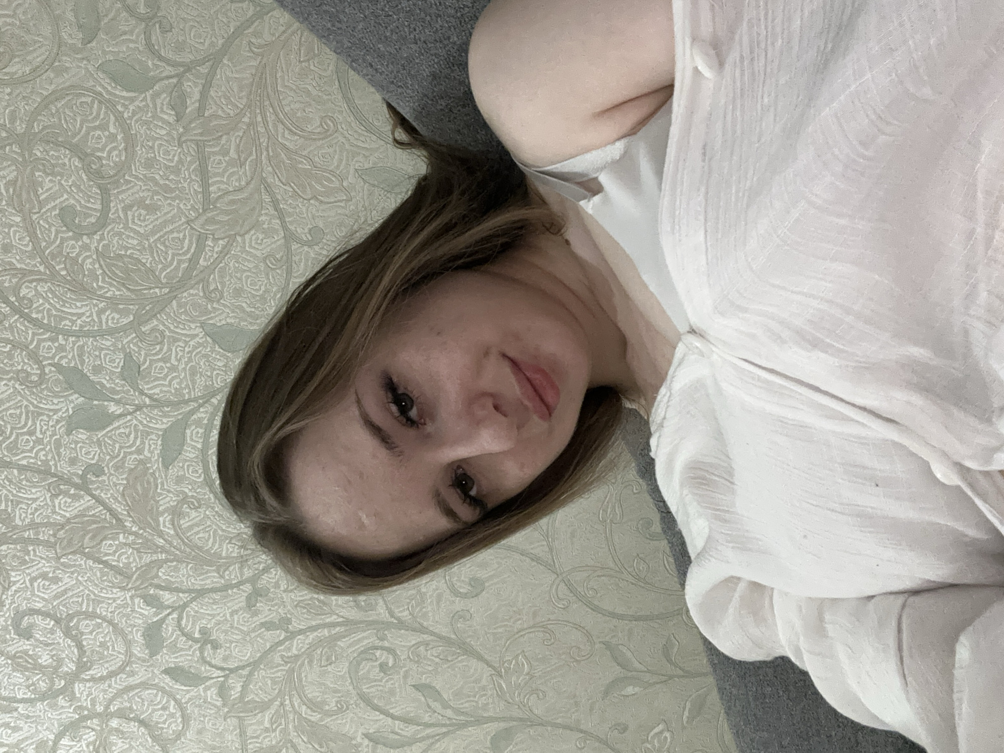 In an indoor setting, a young woman with shoulder-length brown hair is reclining on a gray sofa. She is facing the camera, her head tilted slightly to her left. She is wearing a white top, and her left arm is visible. The background consists of a patterned wallpaper with a floral design in light gray and white tones. The lighting appears to be soft, suggesting an indoor ambient light source.