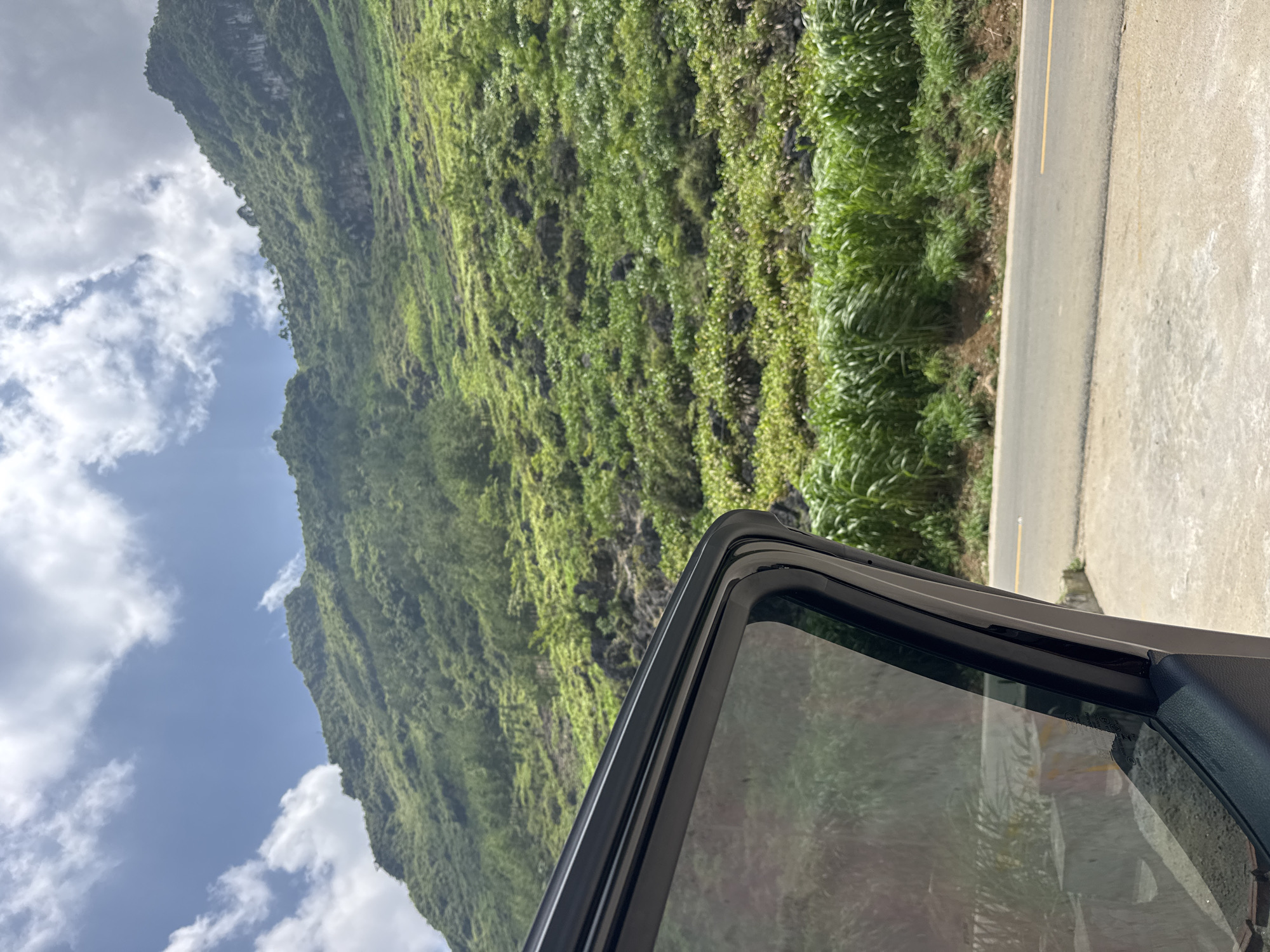 A car door is open, revealing a view of a steep, green, mountainous terrain with a road running alongside it, under a blue sky with white clouds. This scene is in Mèo Vạc Commune, Vietnam.