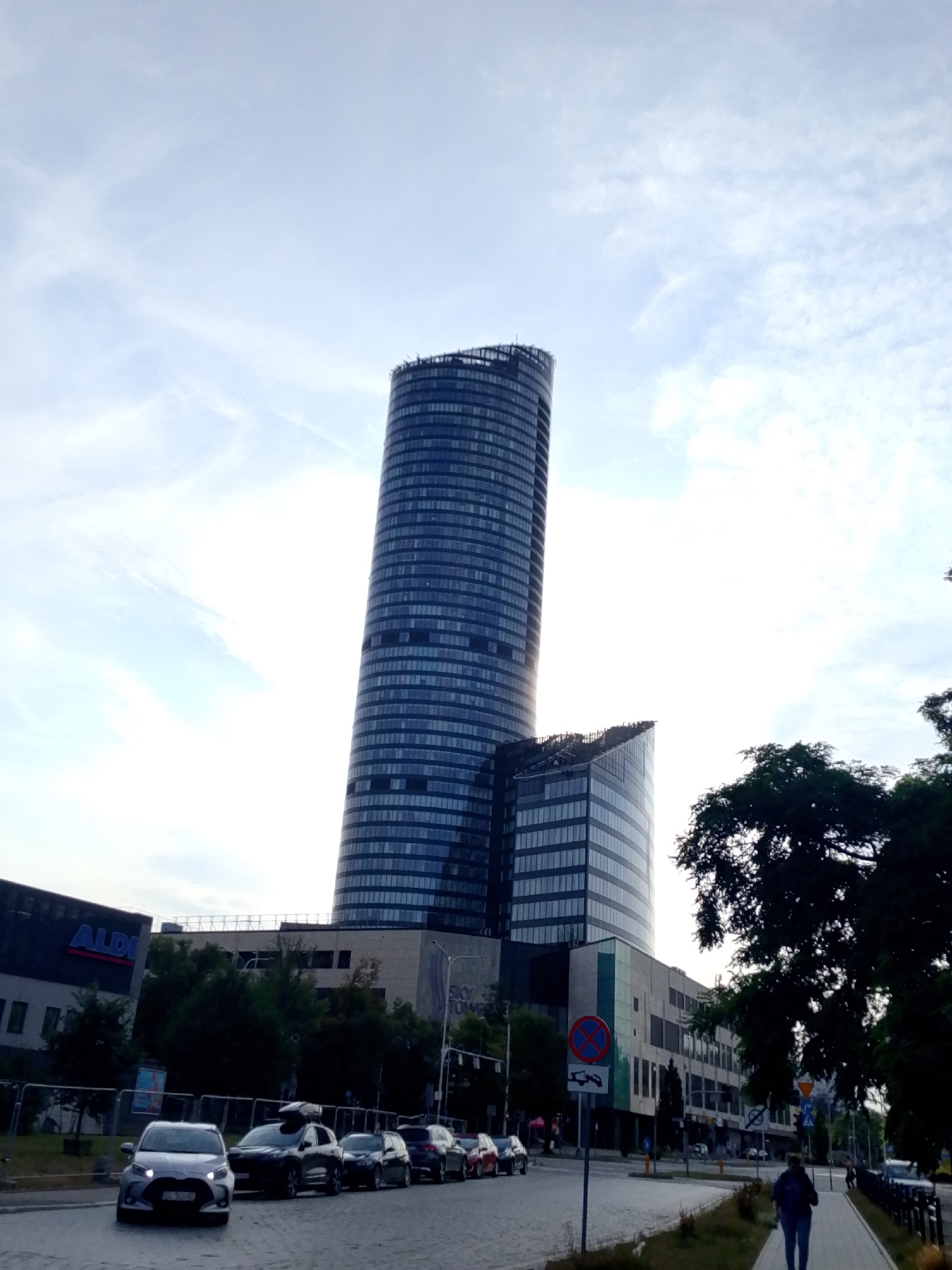 The image shows the Sky Tower in Wrocław, Poland, on a cloudy day. The skyscraper, a prominent landmark in the city, is visible in the background, its glass facade reflecting the sky. In the foreground, a street is lined with cars, some parked and others in motion. A person is walking on the sidewalk, and trees and buildings, including an Aldi store, are also present in the scene. The overall impression is of an urban environment on a somewhat overcast day.