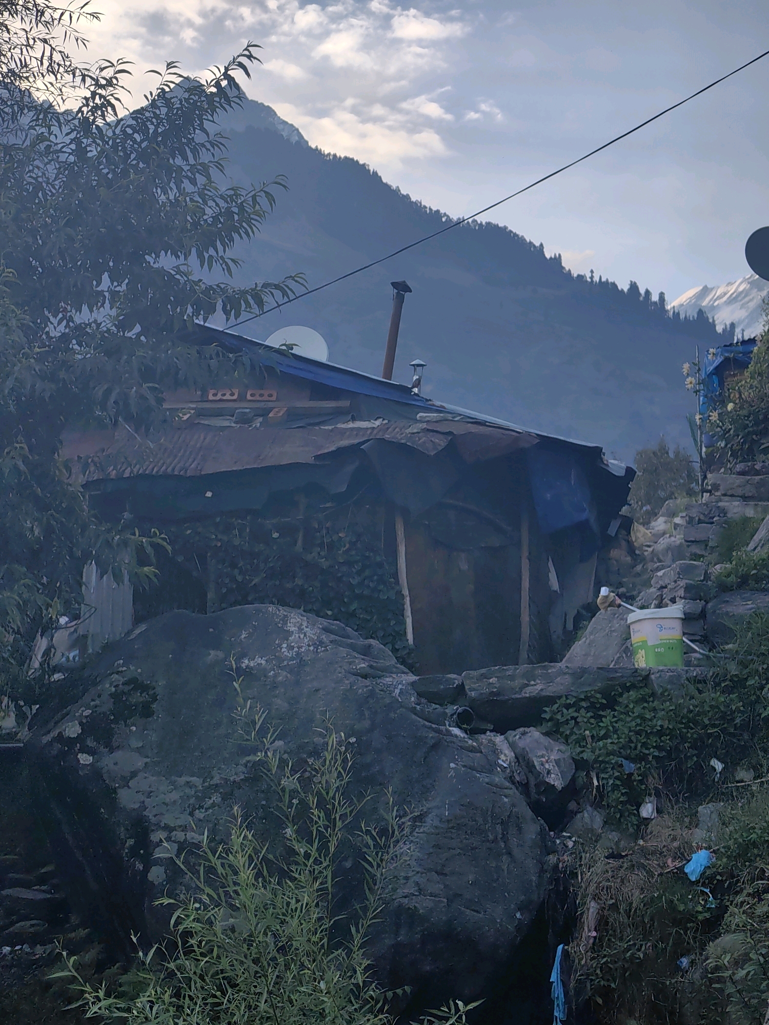 The image depicts a rustic dwelling in Vashisht, India, nestled against a backdrop of majestic mountains. The foreground is dominated by large, dark rocks and lush green vegetation. The dwelling itself appears to be constructed from salvaged materials, including corrugated metal sheeting and a blue tarp. A satellite dish is visible on the roof, suggesting a connection to the outside world. A metal chimney extends from the roof, indicating that the structure is inhabited and likely used for heating.
Further into the scene, the mountainous terrain rises steeply, with a band of trees clinging to the slopes. The sky is partly cloudy, with soft light filtering through the clouds. In the distance, snow-capped peaks are visible, hinting at the elevation and geographical context of Vashisht. The overall impression is one of rugged beauty and a simple, unadorned way of life in a remote, mountainous setting. The presence of a green and white bucket with some writing on it, along with some discarded plastic bags, suggests that the area is lived in and utilized by people, adding a touch of realism to the otherwise serene mountain landscape.