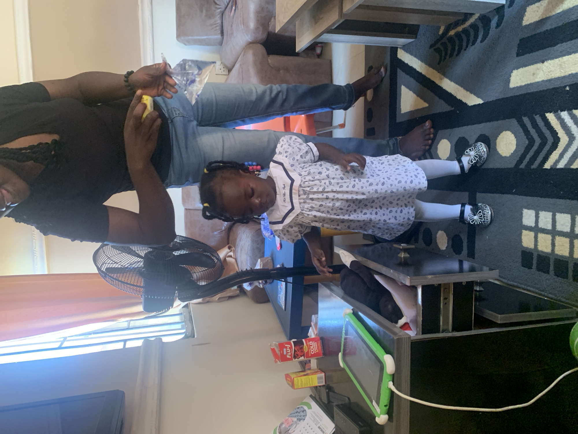 An adult, facing away from the camera and holding a small yellow object in their right hand and a clear plastic bag in their left, stands in an indoor setting. They are wearing a black t-shirt and blue jeans. In front of the adult, a young Black child stands facing the camera. The child has braided hair styled with yellow ties and is wearing a white dress with a blue patterned print, white stockings, and black shoes adorned with bows. The child's gaze is directed downwards.
The environment appears to be a living room or common area. To the left, a black standing fan is visible. On a dark-colored table in the foreground, a green tablet is charging, and several boxes of medication, including "FLU-J Cold, Cough & Paediatric," are present. Behind the table, a dark blue table is situated. In the background, a brown sofa is visible, and to the far right, part of a light-colored wooden table or counter is in frame. The floor is covered with a patterned rug featuring geometric shapes in black, grey, white, and yellow. The lighting suggests a daytime scene, with natural light entering from a window partially obscured by orange curtains to the left. The overall impression is a domestic scene in Gura Top, Nigeria.