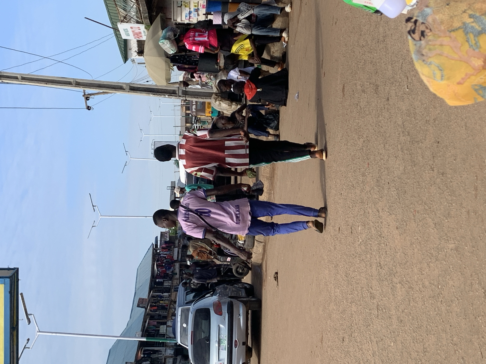 This media file captures a vibrant street scene, likely in Bukuru, Nigeria. The image is oriented vertically, as if the photographer is standing on a street.
The main subjects are a group of people, mostly young men, dressed in casual clothing. One man in the foreground wears a maroon and white striped shirt and dark pants. To his right, another man is seen from the back, wearing a light pink shirt with text and blue pants. Further back, other individuals are visible, some seated, others standing. They appear to be gathered in a market or busy street area.
Objects present include a few motorcycles and a silver car. In the background, market stalls or shops are visible, with awnings and displays of goods. There are also streetlights and utility poles, suggesting an urban or semi-urban environment. A large, light-colored umbrella or canopy is also visible, possibly for shade.
The setting is a paved road, characteristic of a marketplace or busy street. The sky is clear and blue, indicating it is daytime, likely morning or afternoon, with good weather.
There is visible text on a sign hanging from a building that reads "HERBERT POPOOLA LTD". The pink shirt worn by one of the men has text on it, including "ROWAKAS" and a number "20". The license plate on the car reads "BIW-35746".
The overall impression is one of daily life and commerce in a Nigerian town. The people's postures suggest they might be resting, waiting, or engaged in conversation, creating a relaxed yet active atmosphere.