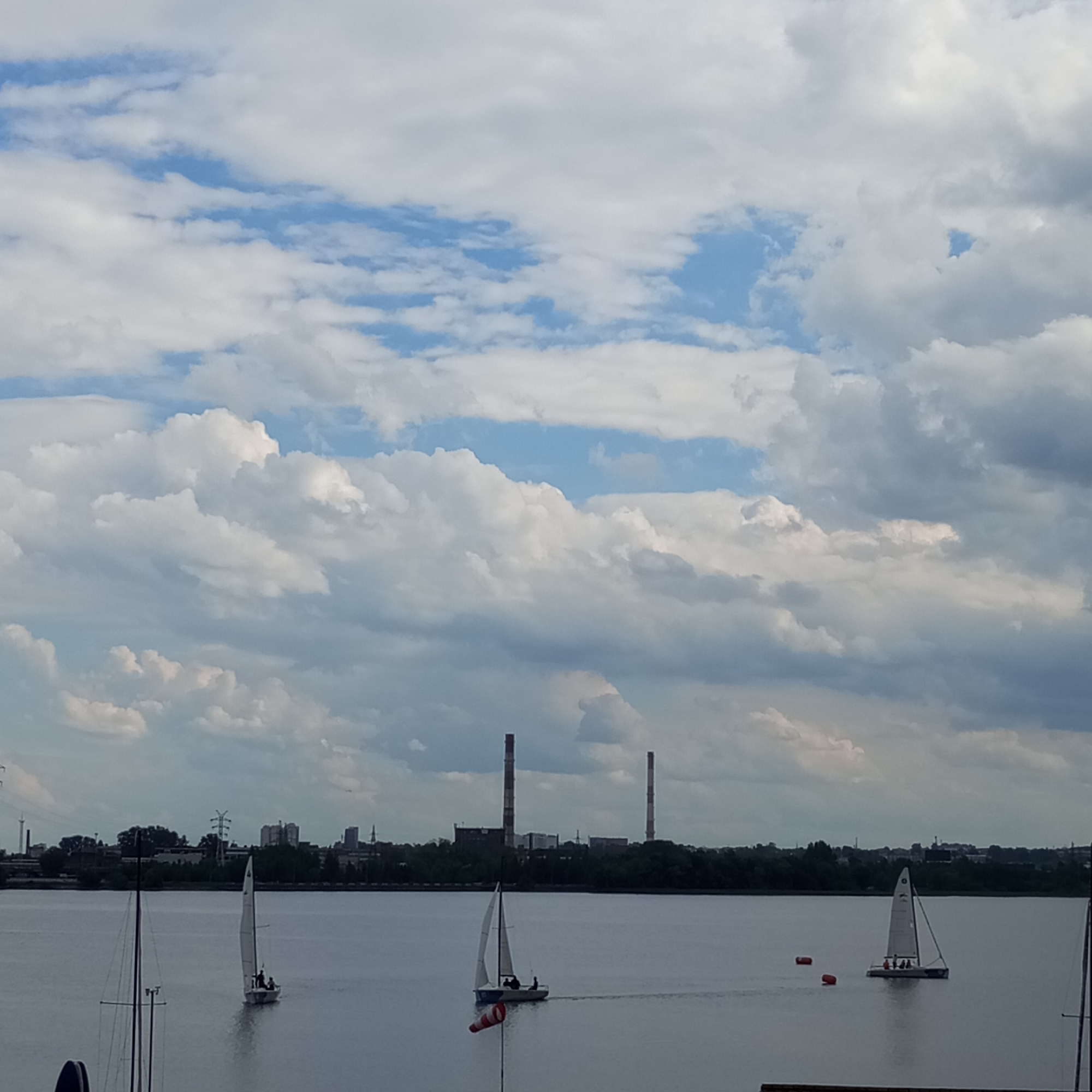 The image depicts a wide expanse of water under a cloudy sky. In the distance, a city skyline is visible with several industrial chimneys. Several sailboats are present on the water, some with occupants. The scene appears to be a recreational area within or near a city, potentially Izhevsk, Russia, given the industrial elements.