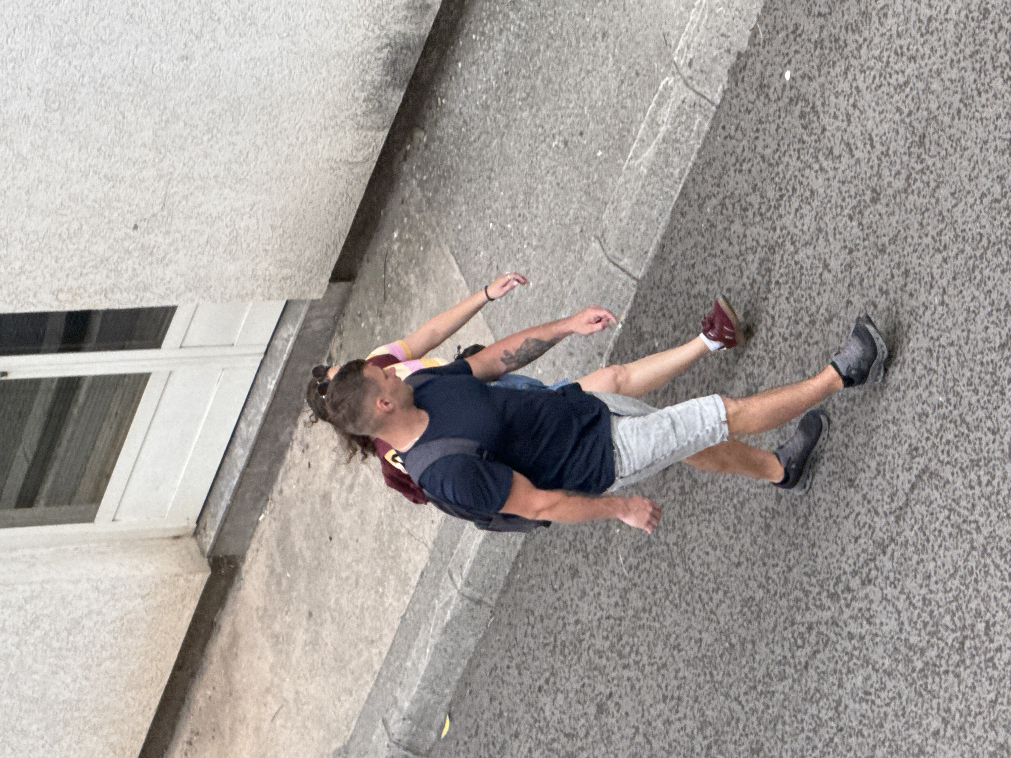 Viewed from an elevated position, a man and a woman walk side-by-side on a concrete sidewalk in Budva, Montenegro. The man, wearing a dark-colored short-sleeved shirt, grey shorts, and dark-colored sneakers, has a backpack slung over his shoulder. Visible tattoos adorn his left arm. The woman, walking to his left, wears a pink and yellow striped top and is barefooted. To the left, the base of a light-colored building is visible, featuring a doorway. The concrete sidewalk extends into the distance.