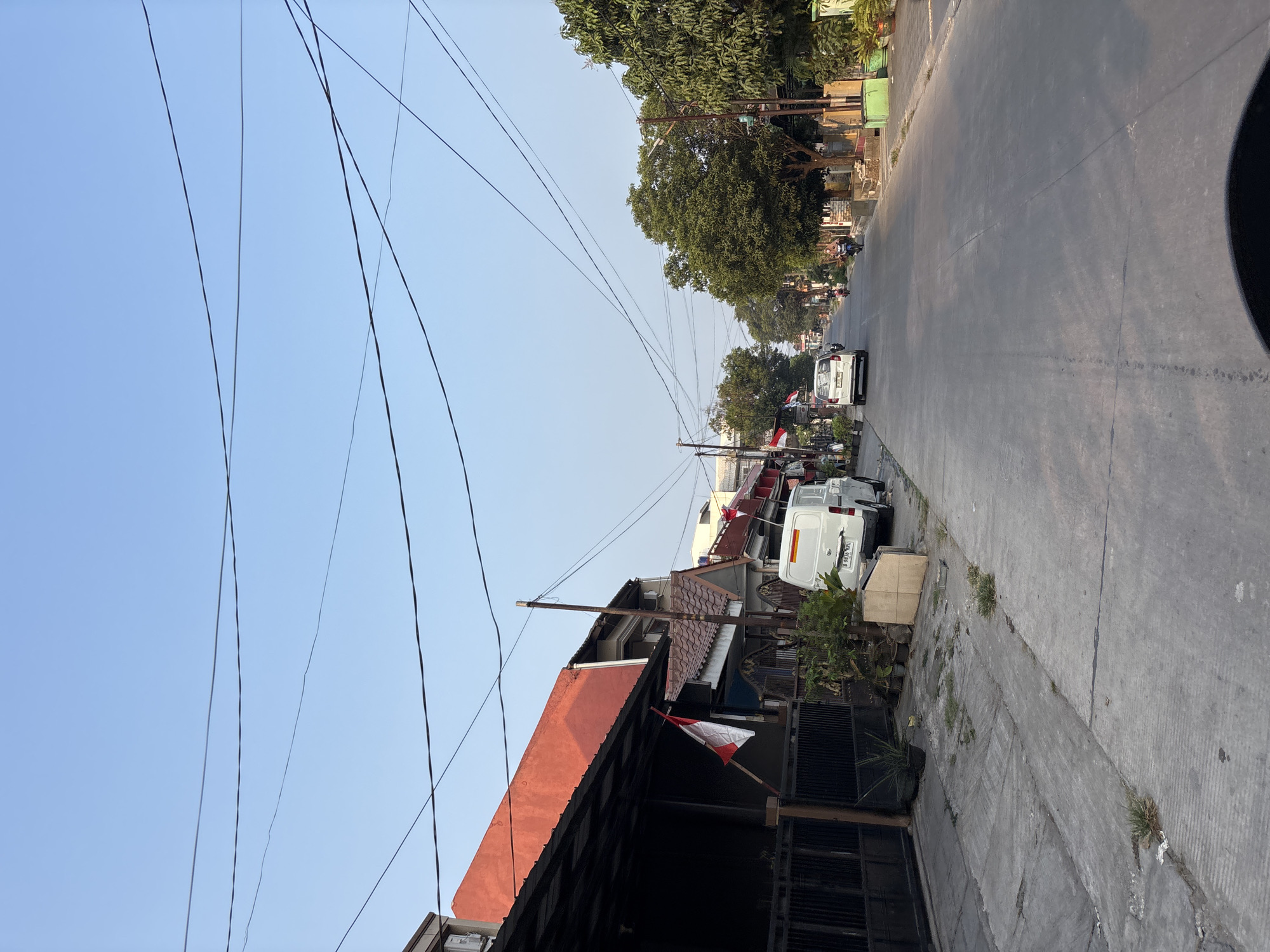 The image depicts a street scene in Bekasi, Indonesia, characterized by residential buildings with red-tiled roofs and several parked vehicles. The road is paved and appears to be a typical urban street, with sidewalks on either side. Overhead power lines crisscross the sky.
On the left side of the street, a row of houses features dark-colored gates and awnings, with potted plants and some greenery visible. Small Indonesian flags are displayed on some of the buildings. A white van with a yellow stripe on its rear is parked near the sidewalk. Further down the street, more houses and trees line the road, and a few motorbikes can be seen.
The sky is clear and blue, indicating fair weather. The lighting suggests it is daytime, likely late afternoon or early morning, given the low angle of the sun casting shadows. The overall atmosphere is that of a quiet residential neighborhood in a city.