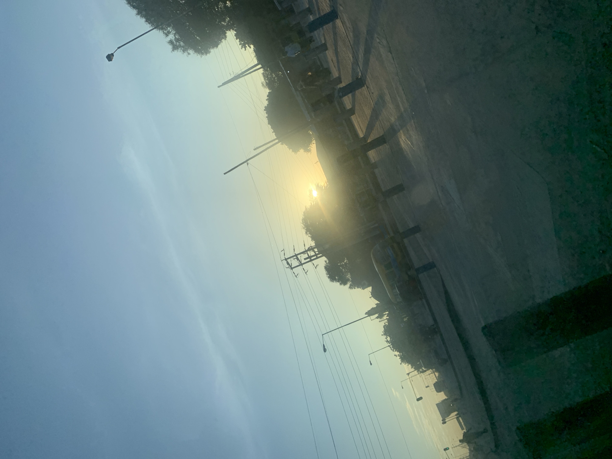 The image shows a street scene likely taken in Dar es Salaam, Tanzania, at or around sunrise. The sun is positioned in the center of the frame, partially obscured by a hazy atmosphere. Utility poles and electrical wires are visible against the sky. Streetlights line the road, which appears to be somewhat deserted. A row of trees is along the roadside. The photo is taken from a low angle, possibly from inside a vehicle, and the lower portion of the frame shows a blurred portion of the vehicle's interior.