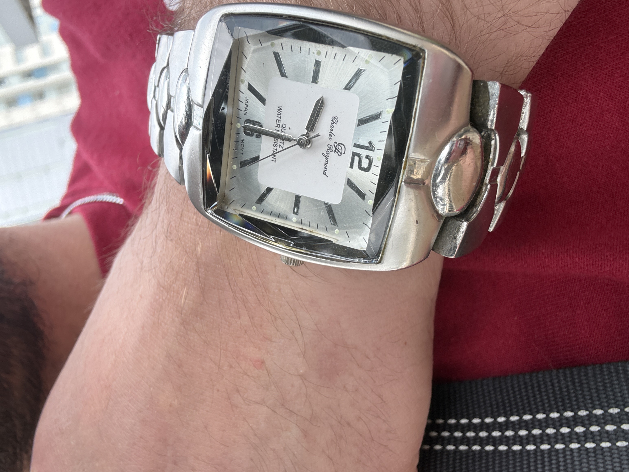 The image presents a close-up shot of a person's hairy forearm and wrist, featuring a prominent silver-toned wristwatch. The watch has a rectangular, somewhat square-shaped case and a broad, metallic link band with decorative oval elements. The watch face is light-colored, likely silver, with dark, rectangular hour markers and black hour, minute, and second hands. A white sticker or tag is affixed to the center of the watch face, partially obscuring some details.
Visible text on the watch face includes "JAPAN MOVT" on the left side, and "WATER RESISTANT" and "QUARTZ" below the center, although these are partially covered by the sticker. The brand name "Charles Raymond" is also visible in script above the center, and the number "12" is prominently displayed at the 3 o'clock position.
The person is wearing a red shirt, visible on the left and upper right portions of the frame. In the background, a blurred, bright scene with light-colored elements suggests an outdoor or window view, possibly of distant buildings, indicating an urban setting. A dark, textured fabric with parallel rows of white stitching, possibly a strap or belt, is visible at the bottom of the image. The lighting indicates it is daytime. The overall image captures the watch and part of the wearer's arm.