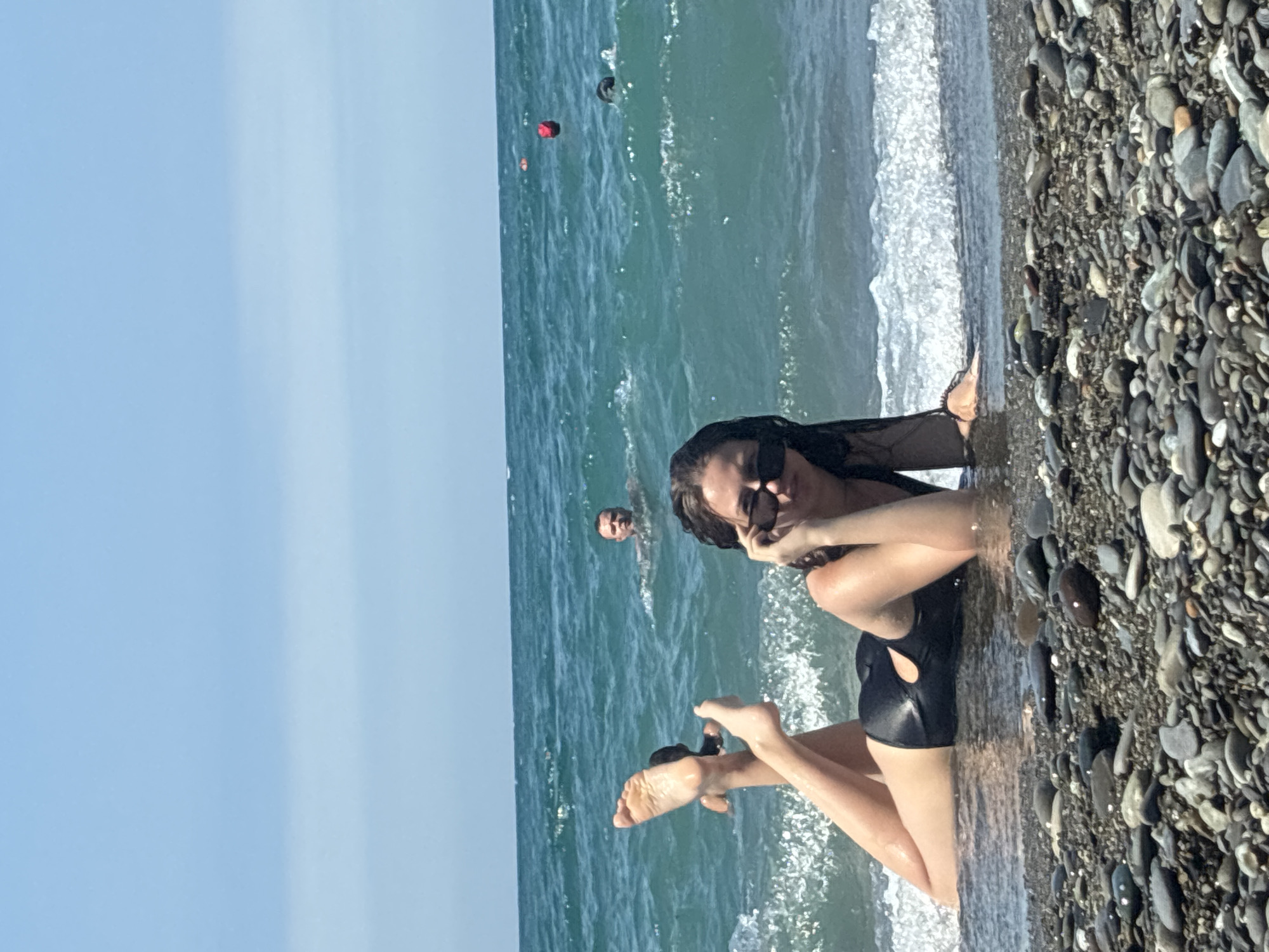 The image captures a sunny day at a pebble beach in Sochi, Russia. In the foreground, a young woman with dark, wet hair and sunglasses is lying on the wet sand at the edge of the turquoise sea, propped up on her elbows. She is wearing a black one-piece swimsuit with a cut-out detail. Her legs are extended and slightly crossed, with her feet in the shallow water. In the background, other people are visible in the water, swimming. One man is swimming closer to the shore, and further out, two buoys are floating in the sea. The sky is clear and blue. The beach is covered with an assortment of smooth pebbles of various sizes and colors.