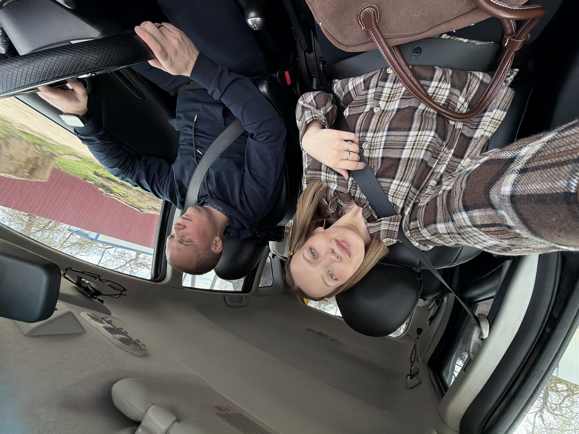 The image shows two people sitting in a car. The man on the left wears a dark jacket and a seatbelt and is holding the grab handle. The woman on the right wears a brown plaid shirt and a seatbelt, and a brown bag is next to her. The car's interior is visible, and the exterior shows a blurred landscape with trees and buildings. Glasses are resting in the overhead console. The location may be in the vicinity of Вельмогово, Russia, based on visual cues from the environment.