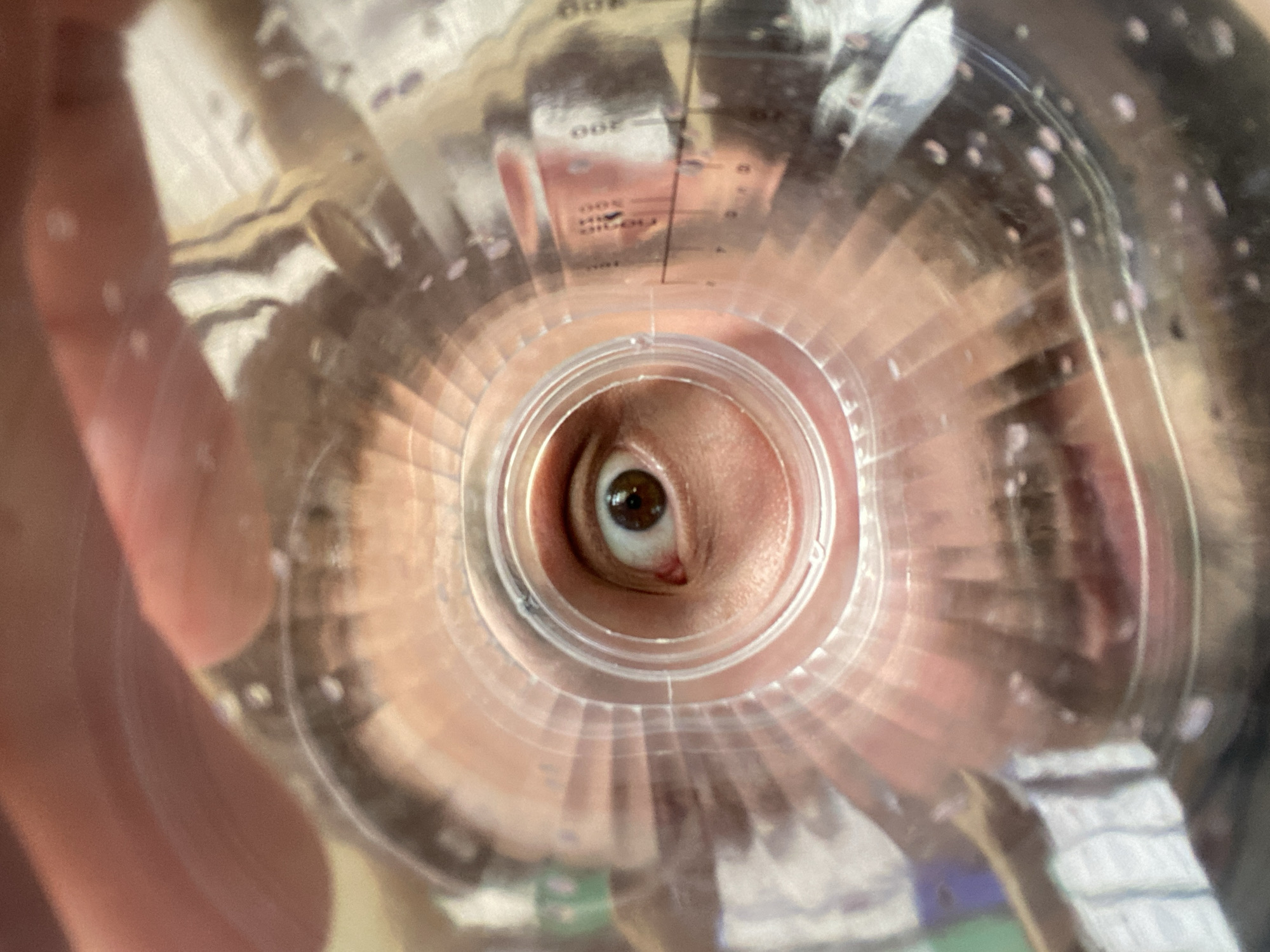 An extreme close-up shot captures a human eye looking through a clear plastic bottle cap. The eye is brown and wide open, with visible eyelashes and eyebrows. The plastic cap has a ribbed edge and appears to be part of a container with volume markings on the side, suggesting it could be a measuring cup or a bottle. The background is blurred and reflects light, with hints of what might be other objects or surfaces. The overall impression is of a distorted and magnified view. There is no indication of the location being Luzhitsy, Russia.