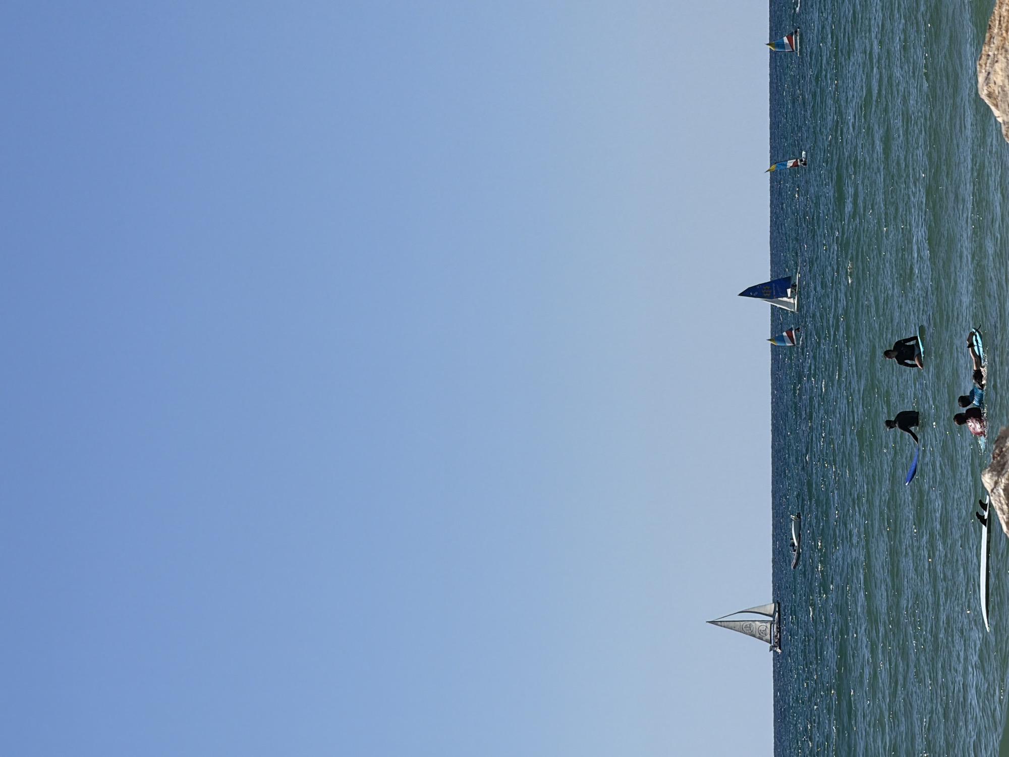 The image shows a sunny day at the beach in Tel Aviv, Israel. The ocean is calm with small waves. Several people are in the water, some are surfing and others are on paddleboards. There are also a few small sailboats with colorful sails. The water is a deep blue, and the sky is clear and a light blue. The sun is shining brightly, casting reflections on the water. In the foreground, there is a rocky embankment. The scene suggests a pleasant day for water activities.