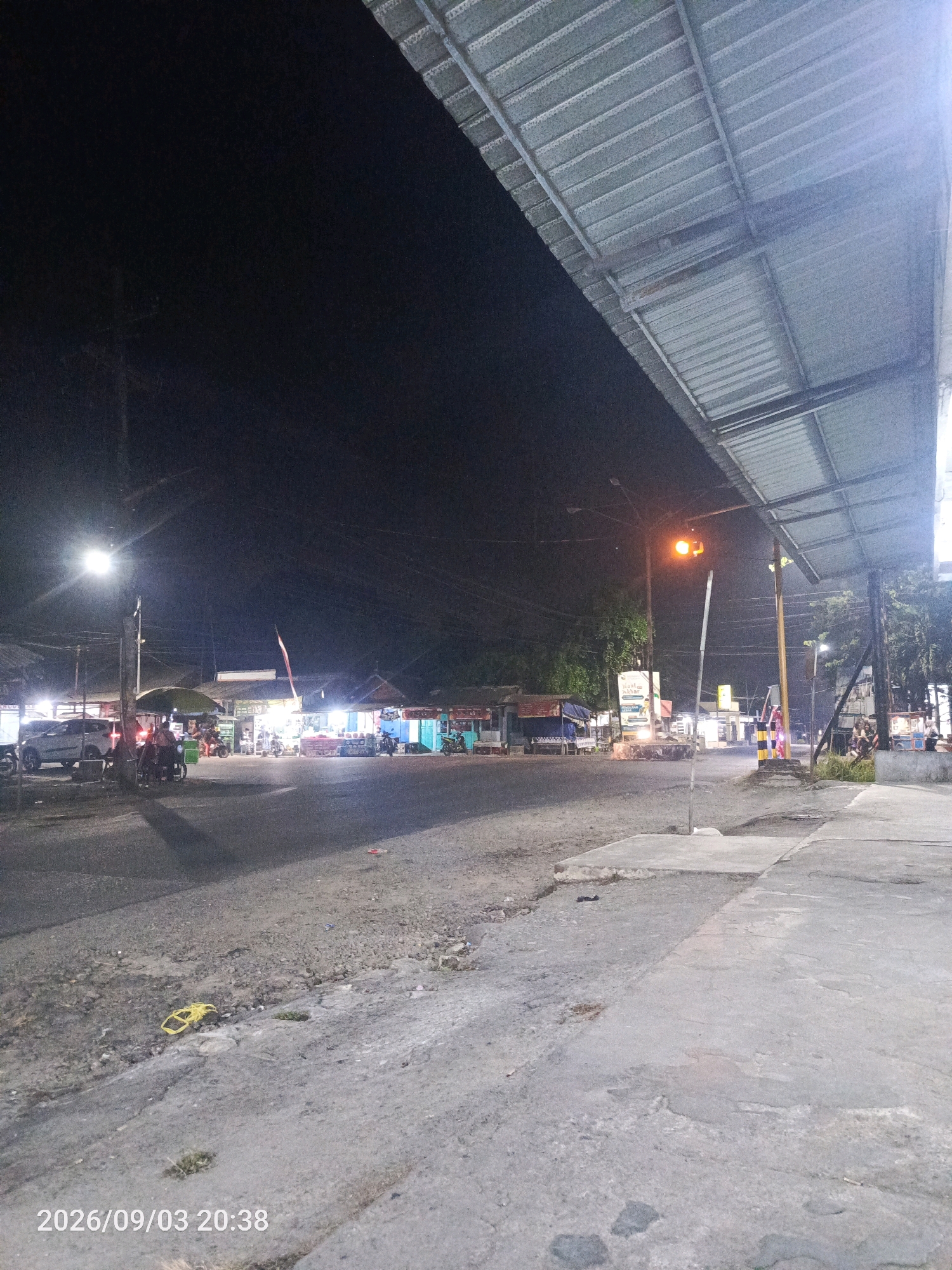 The image depicts a nighttime street scene in Kesilir, Indonesia. A corrugated metal roof structure extends from the right side of the frame, casting a shadow. The street is paved and appears to be a commercial area with various stalls and shops illuminated by artificial lights. Several vehicles, including cars and motorcycles, are parked or in motion. A street lamp illuminates the intersection. In the foreground, a yellow plastic bag lies on the ground. A timestamp in the lower left corner reads "2026/09/03 20:38". The overall atmosphere is that of a busy roadside market at night.