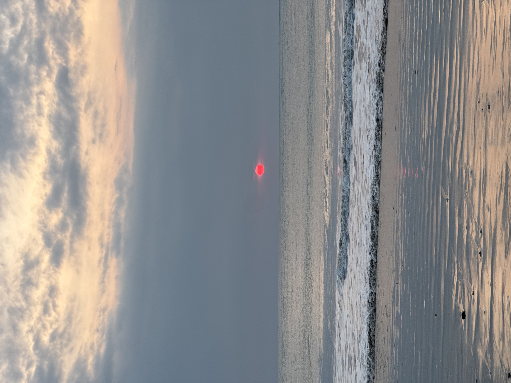 The image depicts a serene oceanfront scene in Kuta Selatan, Indonesia. A vibrant red sun is partially obscured by clouds, casting a warm glow on the horizon. The sky is a gradient of soft peach and grey hues, with scattered clouds adding texture. Gentle waves roll onto a sandy beach, their foamy crests creating a delicate pattern. The wet sand reflects the subdued colors of the sky, creating a mirror-like surface. In the distance, a tiny boat can be seen on the vast expanse of the ocean. The overall atmosphere is tranquil and picturesque, characteristic of a sunset by the coast.