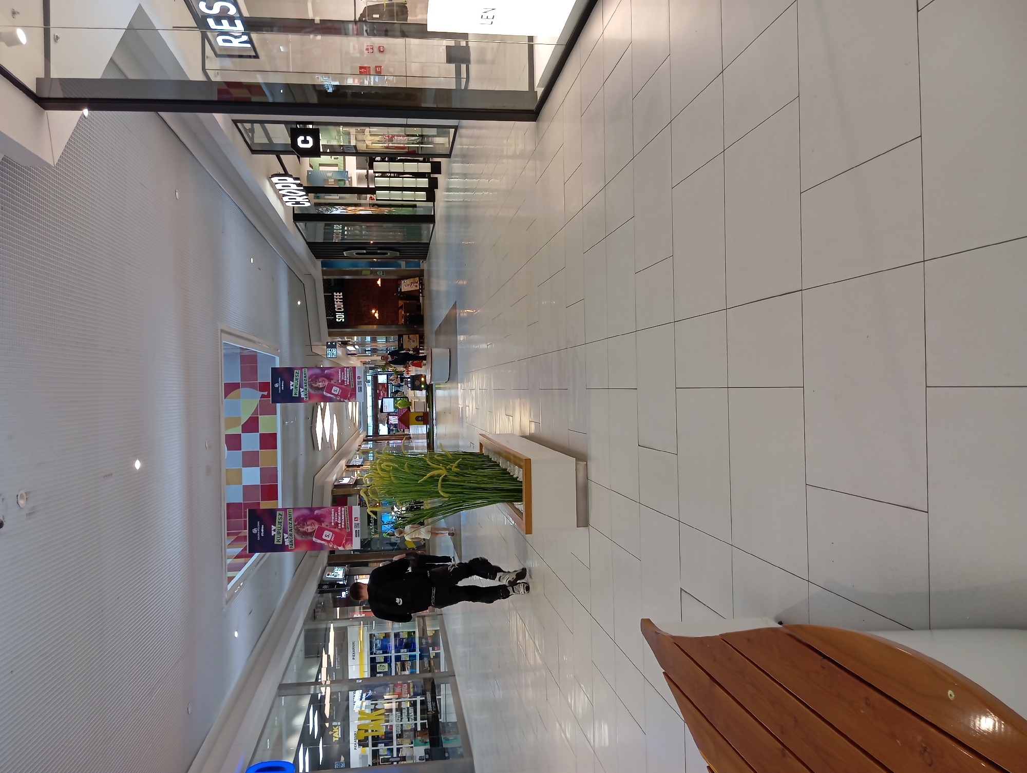 The image shows the interior of a shopping mall with bright white tiled floors and a high, white, corrugated ceiling. A man in black pants and a black shirt, wearing roller skates, is walking away from the camera. He is carrying a black backpack. To his left, a rectangular planter filled with tall, green artificial plants sits on the floor. In the background, several shopfronts are visible. One sign reads "RE." Another sign has the word "CROPP" in large letters. Further down the corridor, a shop called "SOI COFFEE" is advertised. To the left, banners hang from the ceiling, one with the text "TV NAGRADZA!" The overall impression is that of a modern, well-lit shopping environment. The location is the CAMPORE Shopping Centre in Wołomin, Poland.