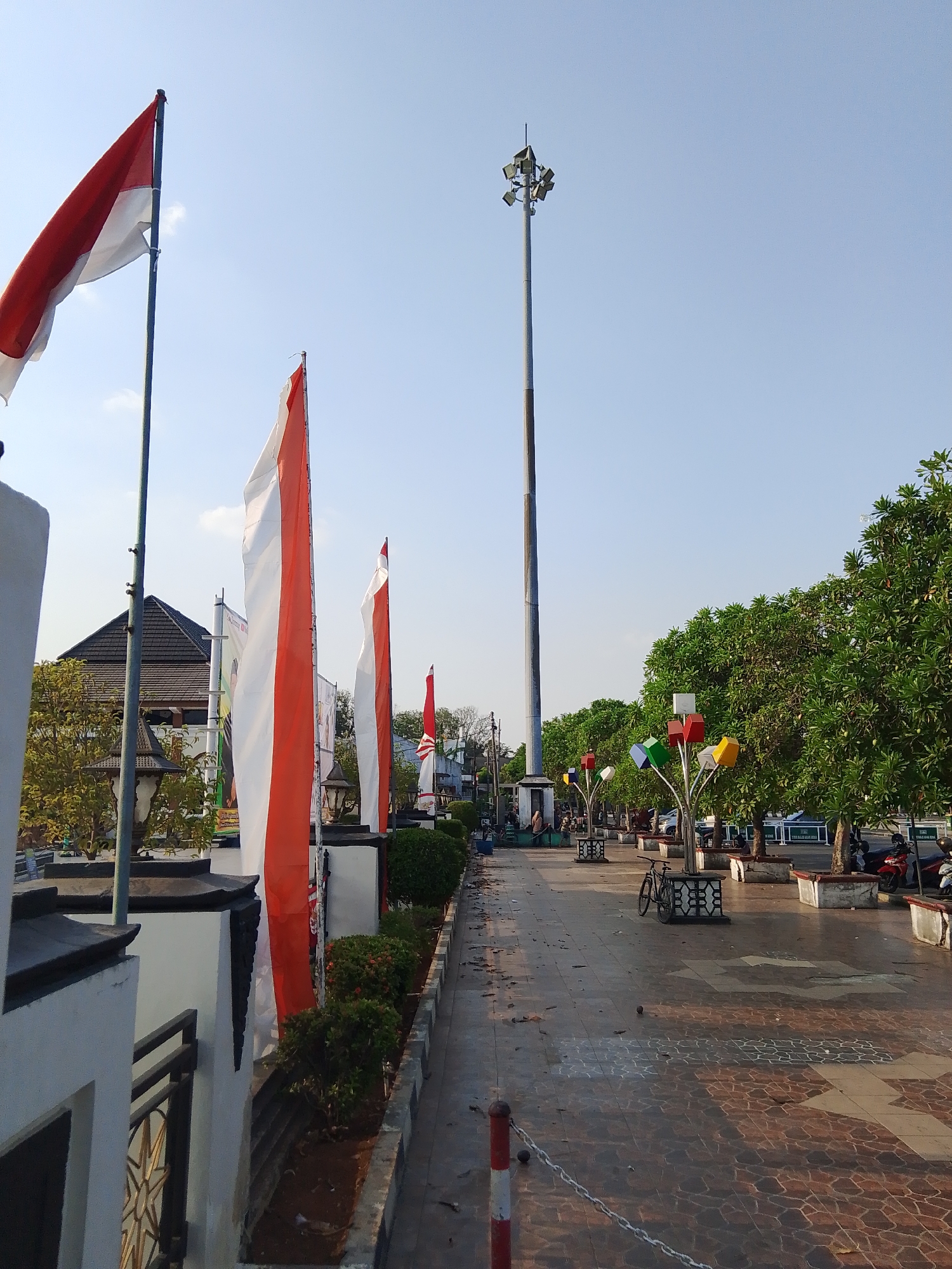 This image depicts a public square in Demak, Indonesia. The square features a large flagpole with the Indonesian flag prominently displayed, along with several other Indonesian flags lining a walkway. The walkway is paved with decorative tiles and flanked by benches and trees. In the background, a tall light tower stands against a clear blue sky. The scene appears to be a peaceful public space, possibly a park or a town square, with no people actively engaged in the frame, though some individuals are visible in the distance.