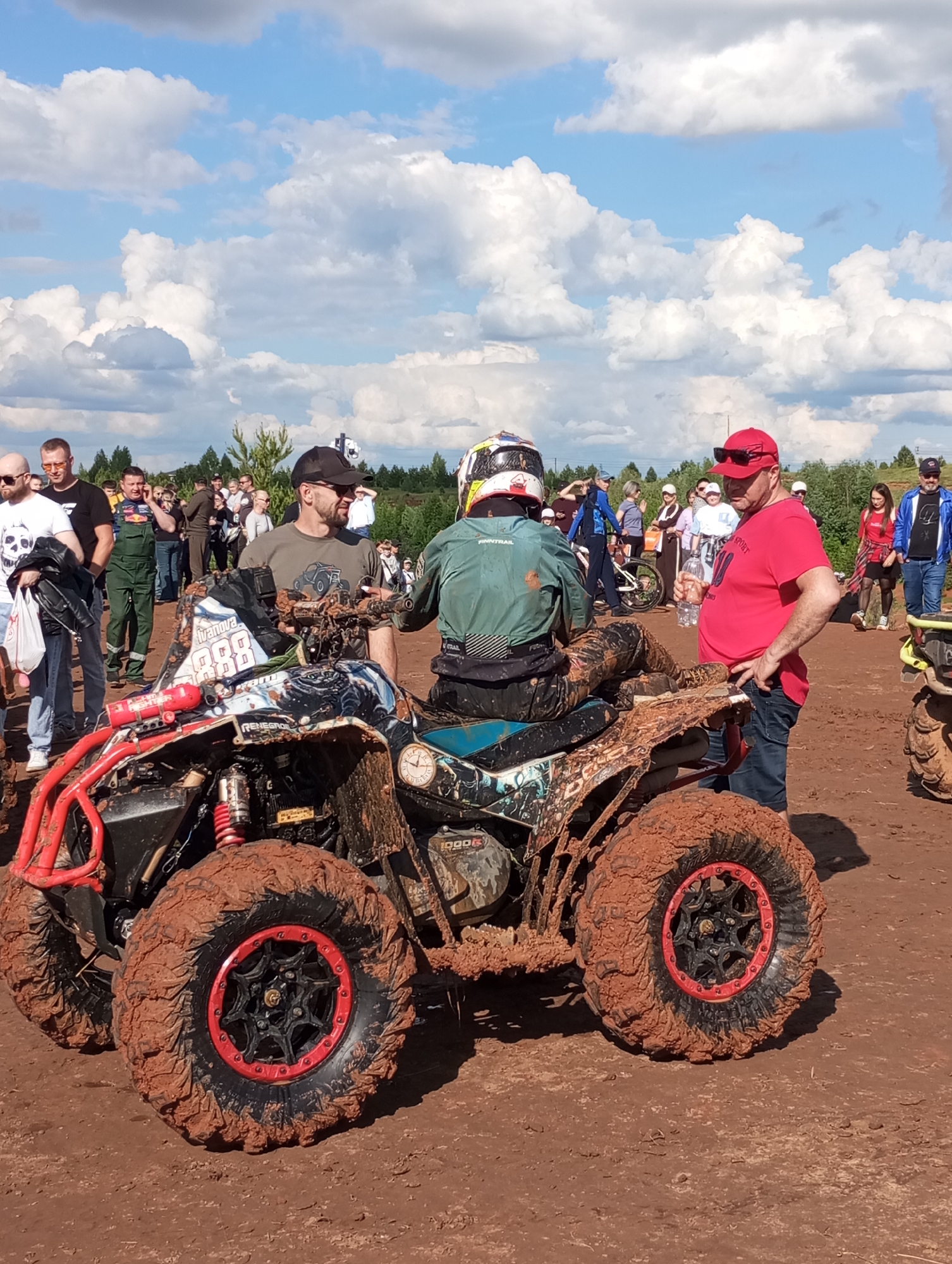 A muddy quad bike is the focal point of an outdoor scene, likely in Izhevsk, Russia. The ATV is covered in red mud, with red accents on its wheels and frame. A person wearing a helmet and green jacket is seated on the vehicle. Two men are standing nearby. The man on the left, wearing a hat and gray-green t-shirt, is looking toward the quad bike. To the right, a man in a red shirt and cap leans against the vehicle, holding a water bottle. Other people are visible in the background, and some are standing and watching. The environment consists of a field covered with red mud and scattered people, with trees and a cloudy sky visible in the distance.