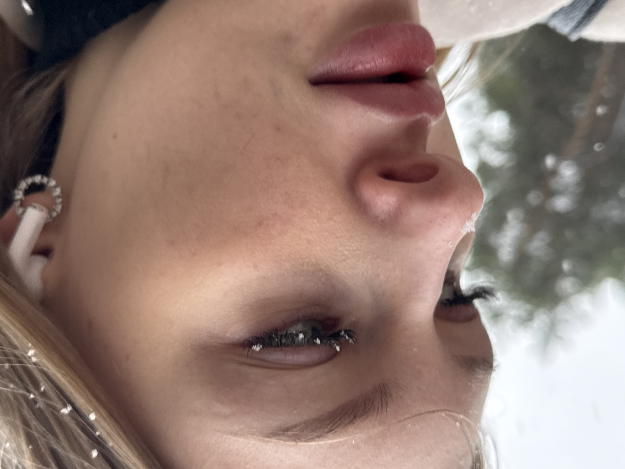 A close-up, low-angle shot shows a woman's face covered with snowflakes. The woman has fair skin, prominent lips, and noticeable dark eyelashes coated with snow. Snowflakes are also visible on her nose, hair, and eyebrows. An earring is visible in her ear, and an electronic device is partially visible. The background, out of focus, appears to show trees and a snowy sky. The location is presumed to be Perm, Russia.