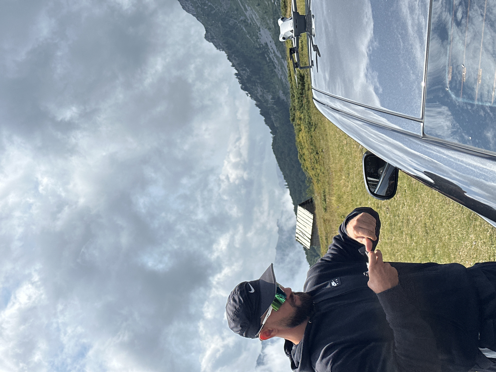 In the city of Katun Vulića, Montenegro, a man wearing a black Nike baseball cap and black hoodie stands near a black car. He has a beard and is wearing white and green sunglasses. He is holding a small black object in his hands and appears to be examining it. A white drone is visible on the roof of the car. The background consists of a grassy field, a small wooden structure with a thatched roof, and a mountain range under a cloudy sky.