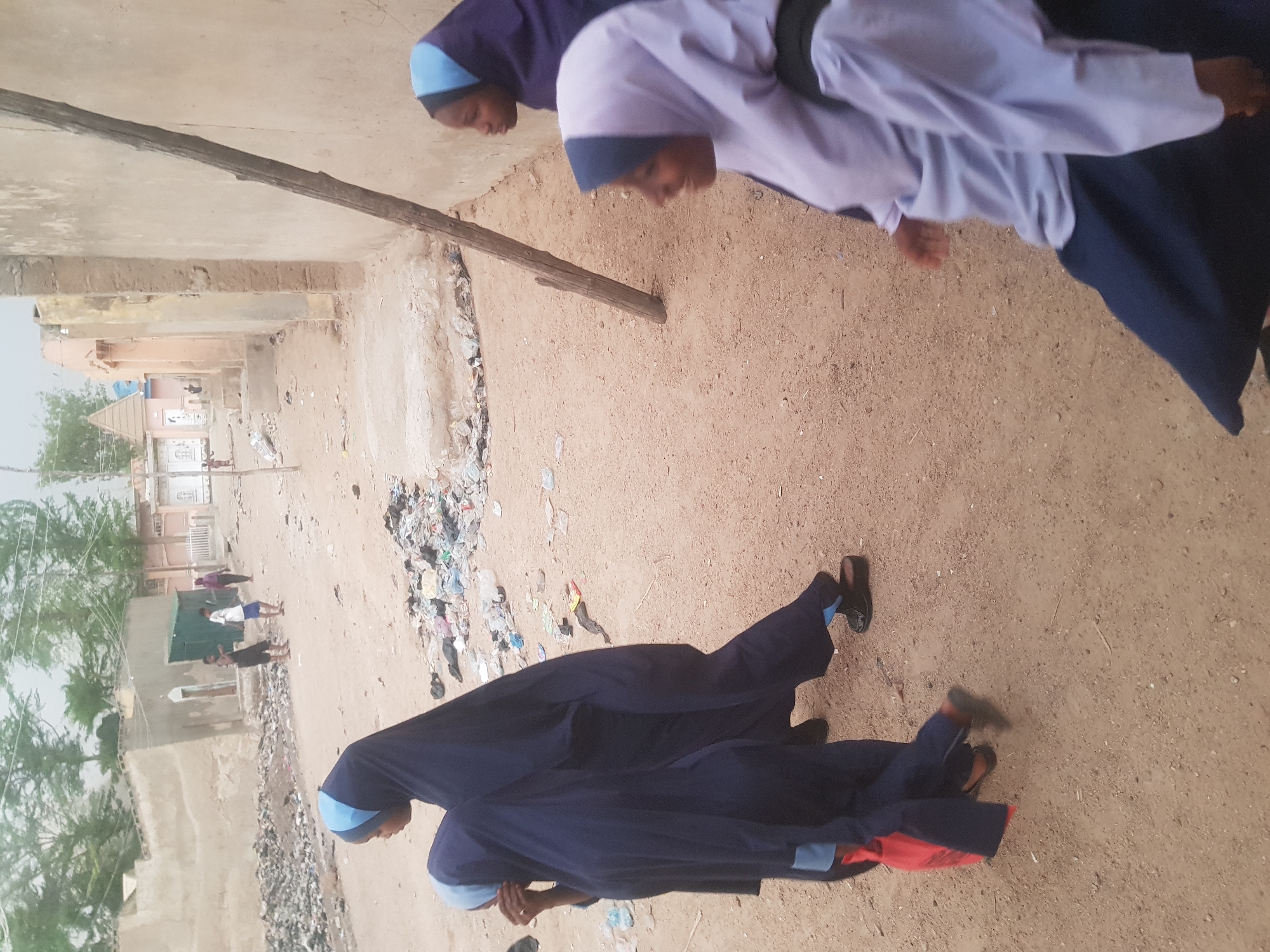 The image shows a street scene in Dutsinma A, Nigeria. In the foreground, two people wearing navy blue robes and light blue head coverings are walking on a sandy surface. The person in the front also has a red object near their feet. Further back, other people are walking on a dirt road, with buildings and trees visible in the background. There is also trash and debris scattered along the road.