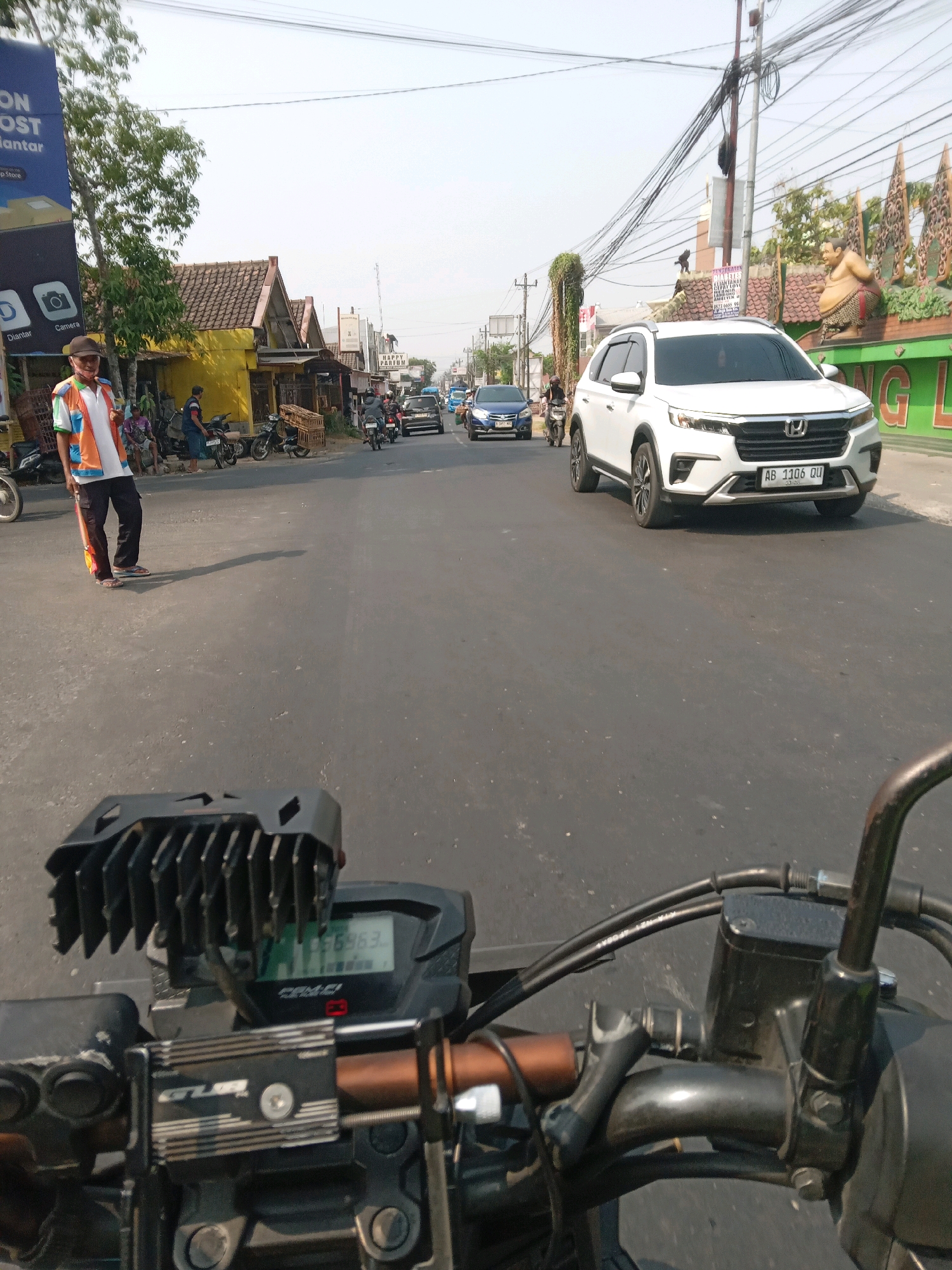 The image depicts a street scene in Banguntapan, Indonesia, taken from the perspective of a motorcycle. In the foreground, the handlebars and digital display of the motorcycle are visible. The display shows the number "656963". The road is paved and appears to be a two-lane street with moderate traffic.
On the left side of the road, a person wearing a reflective vest with orange and white colors, dark pants, and sandals stands on the sidewalk, casting a shadow. Behind this person, there are several parked motorcycles and small shops with yellow and brown facades. A large blue billboard is also present on the left.
The road is occupied by several vehicles, including a white SUV with license plate "AB 1106 QU" in the right lane, and a blue car in the left lane. Other motorcycles and cars are visible further down the road, suggesting a busy urban environment. The background features buildings, trees, and utility poles with a complex network of wires overhead. The sky is clear and bright, indicating daytime. On the right side of the road, there is a decorative wall with a large, rotund statue, and green signage partially visible. The overall scene is characteristic of a commercial or residential street in a Southeast Asian city.