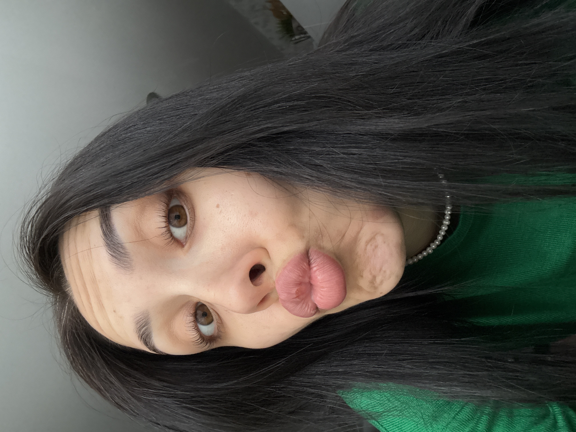 A woman is depicted in a selfie-style, angled shot. She has long, dark hair and brown eyes. She is wearing a green top and a pearl necklace. Her lips are pursed. The background is a blurred neutral tone. The image provides no information suggesting a location in the city of Votkinsk, Russia.