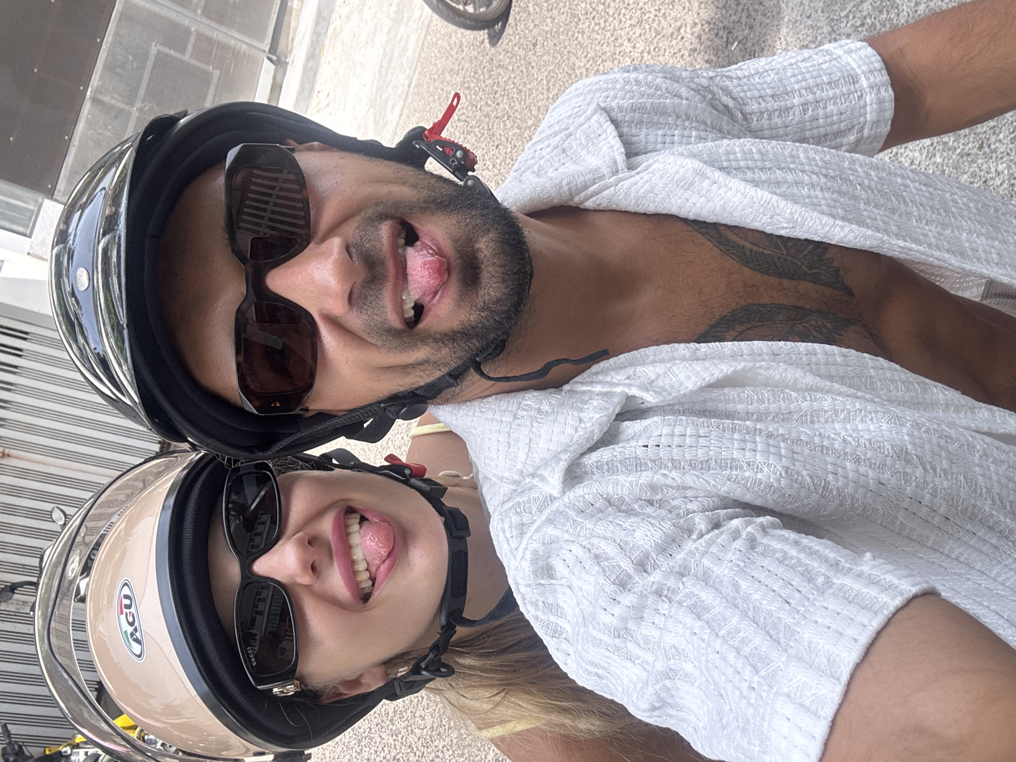 A man and a woman are riding on a motorcycle in Nha Trang Ward, Vietnam. The woman is wearing a beige helmet and sunglasses. She has blonde hair and is sticking her tongue out. The man is wearing a black helmet and sunglasses. He is also sticking his tongue out. Both are wearing white shirts. The background shows a building with striped walls and a paved ground.