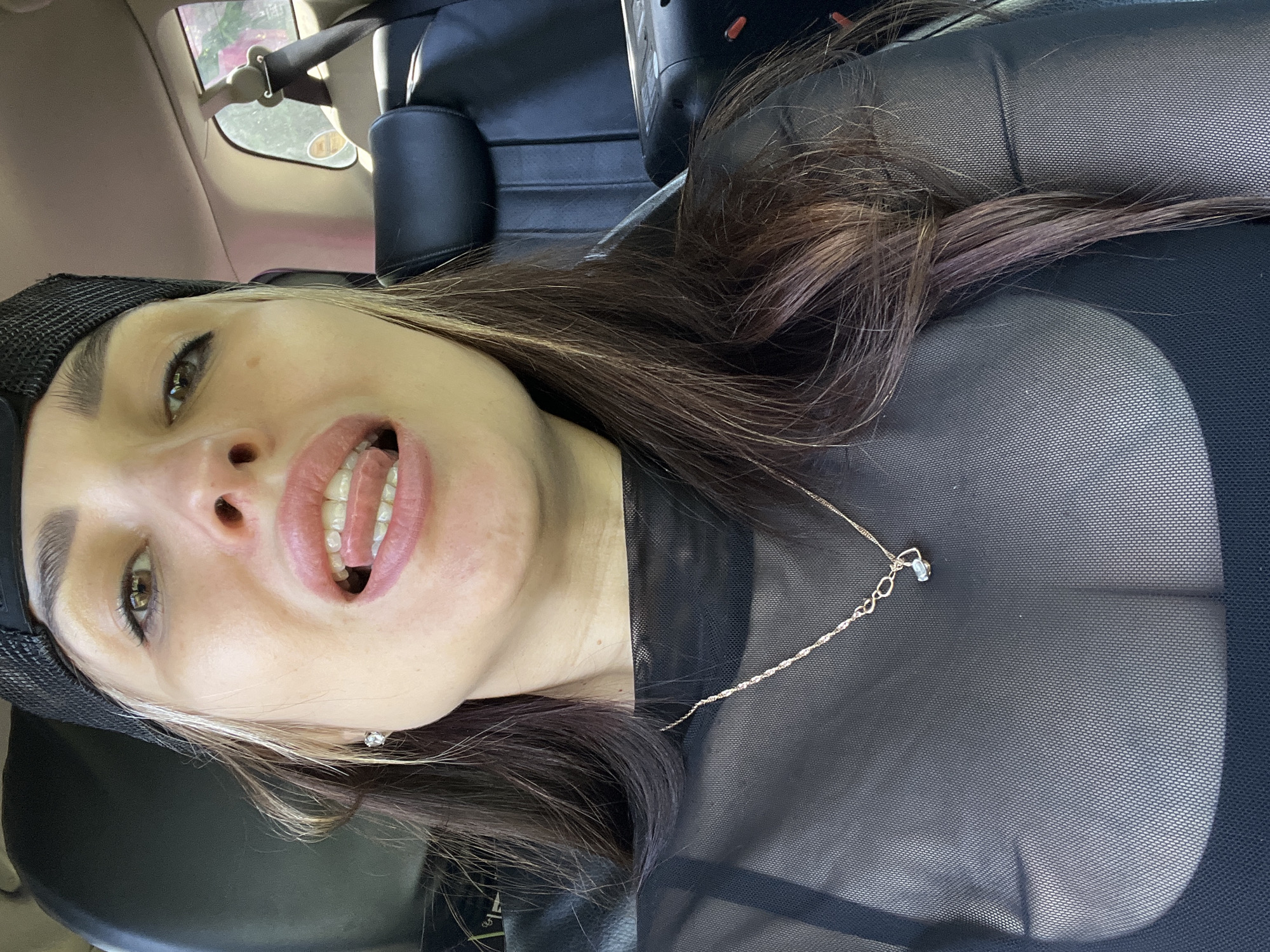 A woman is reclining in the passenger seat of a car. She is wearing a black mesh top and a black baseball cap. She has long brown hair with a streak of blonde. She has a necklace on. She is looking up at the camera with her mouth open, revealing her teeth. The location appears to be Unknown, Unknown.