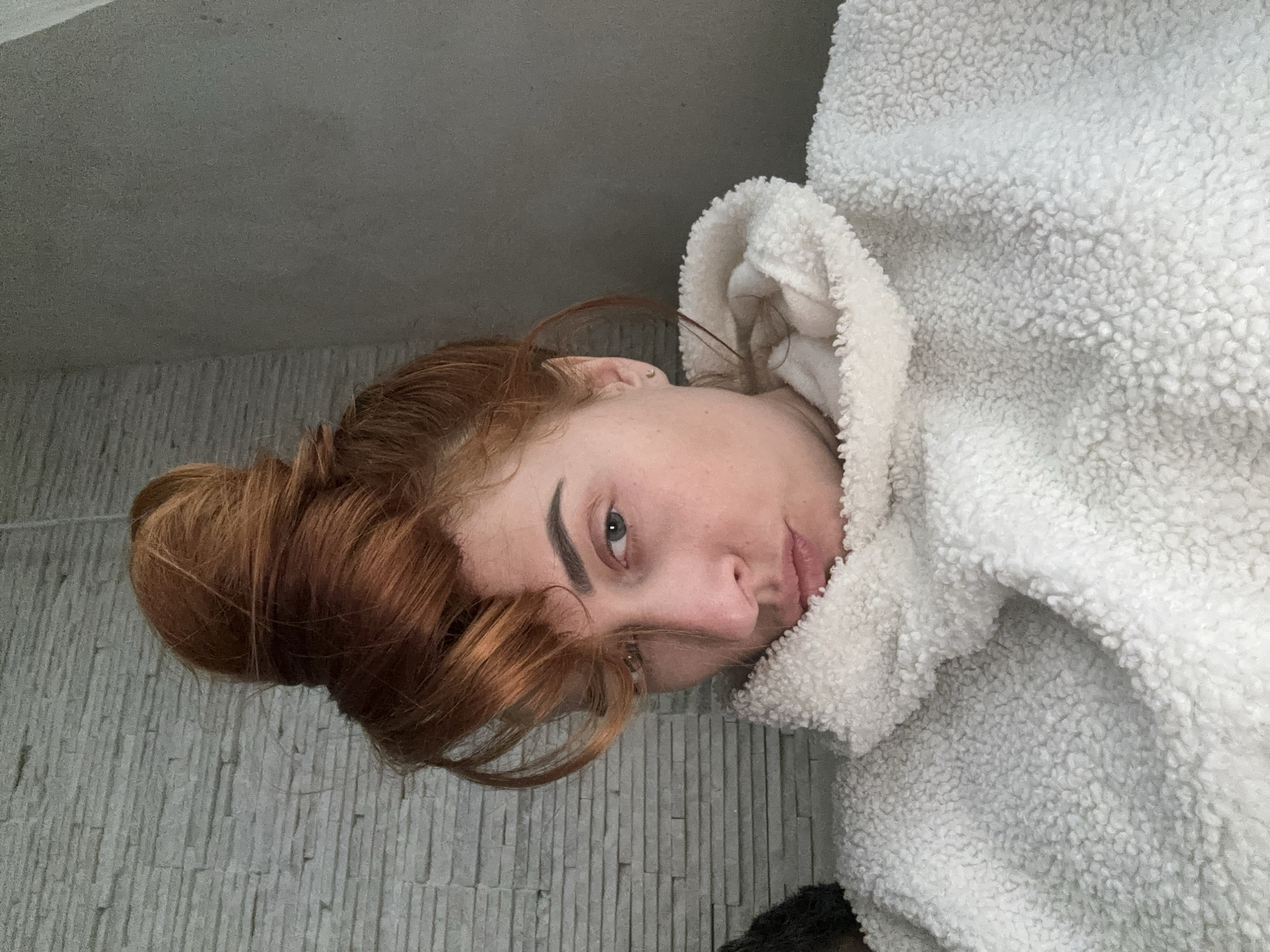 A woman with red hair pulled into a bun is wearing a white fluffy hooded jacket. She is looking at the camera. The background appears to be a stone-like wall and a ceiling. The image's orientation is rotated counter-clockwise. The location in the city of Budva, Montenegro is not discernible from the image.