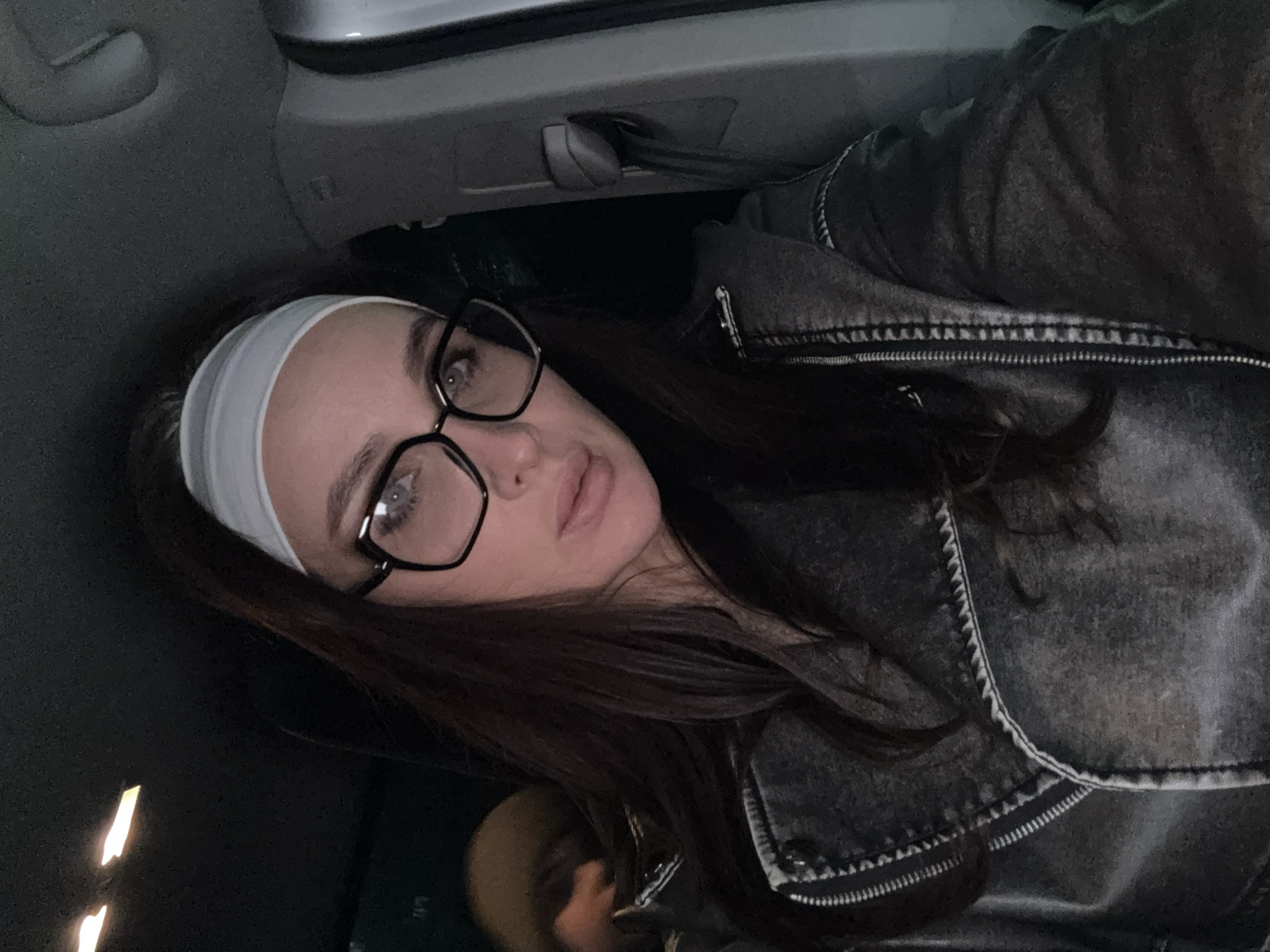 A woman with long dark hair and a white headband is wearing black framed glasses and a dark gray jacket. She is sitting in a car. The interior of the car is visible in the background. The photograph was taken indoors.