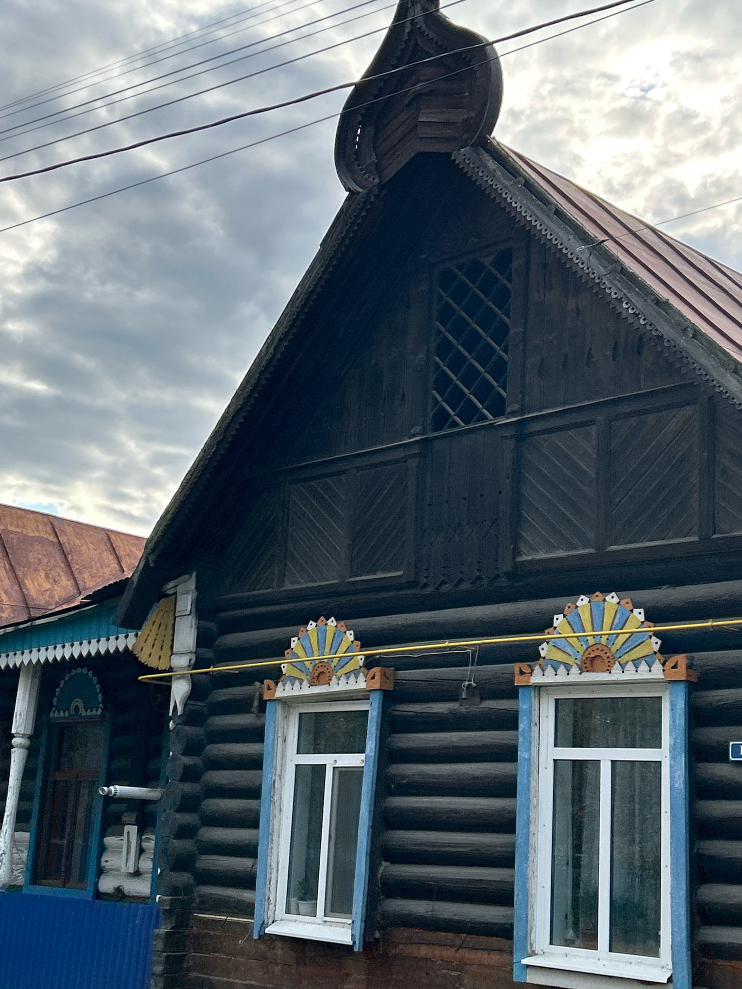 A low-angle, close-up shot captures the upper facade of a dark wooden log house, likely in the city of Tomsk, Russia, given the traditional architectural elements. The house features a steeply pitched, dark wooden gable roof with decorative wooden trim along the eaves. A diamond-shaped lattice window is visible in the upper gable. Below the gable, the log walls are visible, with two white-framed windows featuring colorful, fan-shaped decorative elements above them. A yellow pipe runs horizontally across the facade, just above the windows. To the left, part of an adjacent building with a reddish-brown roof and blue trim is visible. The sky is overcast with dramatic clouds, and power lines are strung across the upper portion of the frame. No people are present in the scene.