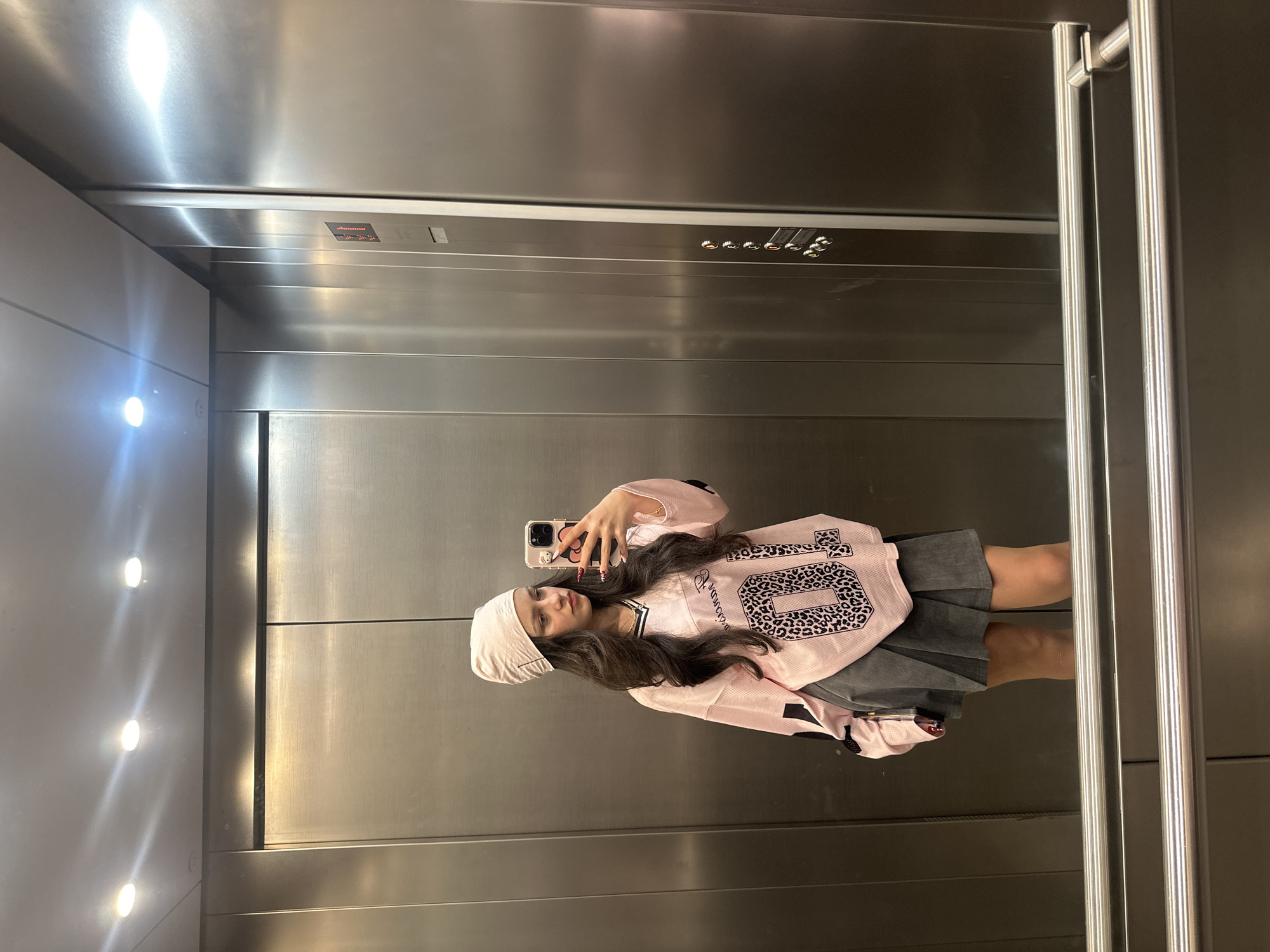 A young woman is taking a selfie in an elevator. She is wearing a pink and black jersey with the number "04" printed on it, a gray skirt, and a white beanie. The elevator has stainless steel walls and a row of lights on the left side. There are also elevator controls on the top of the back wall. There is no clear evidence of the location being in Yerevan, Armenia.