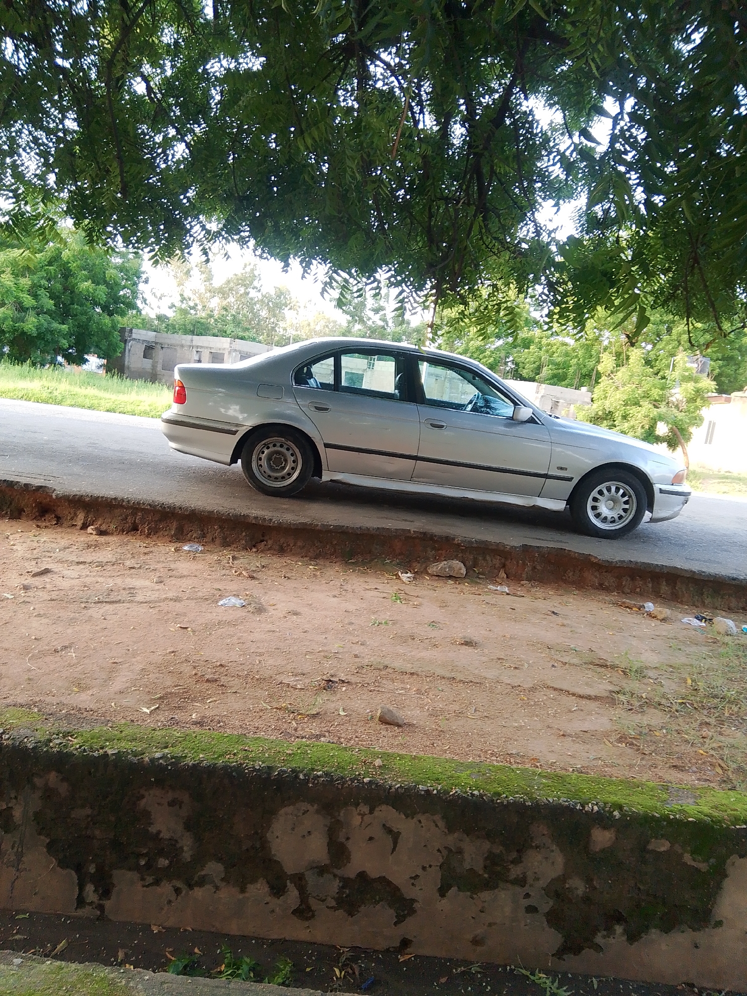 A silver sedan is parked on the side of a paved road, with its tires on the asphalt. The car is viewed from the side, with the rear half of the vehicle more prominent. The car has tinted windows and a black trim along the bottom of the doors.
The road appears to be in a somewhat rural or less developed area, as there is a dirt embankment next to the road, with some scattered debris. Beyond the embankment, there is lush green vegetation and trees. In the background, there are buildings, one of which appears to be unfinished or under construction.
The scene is bathed in natural light, suggesting it is daytime. The abundance of green foliage and the presence of scattered debris on the ground could indicate a warm or humid climate. The overall impression is a quiet, everyday scene in a semi-urban or rural setting in Jalingo, Nigeria. No people are visible in the image, and there is no visible text.