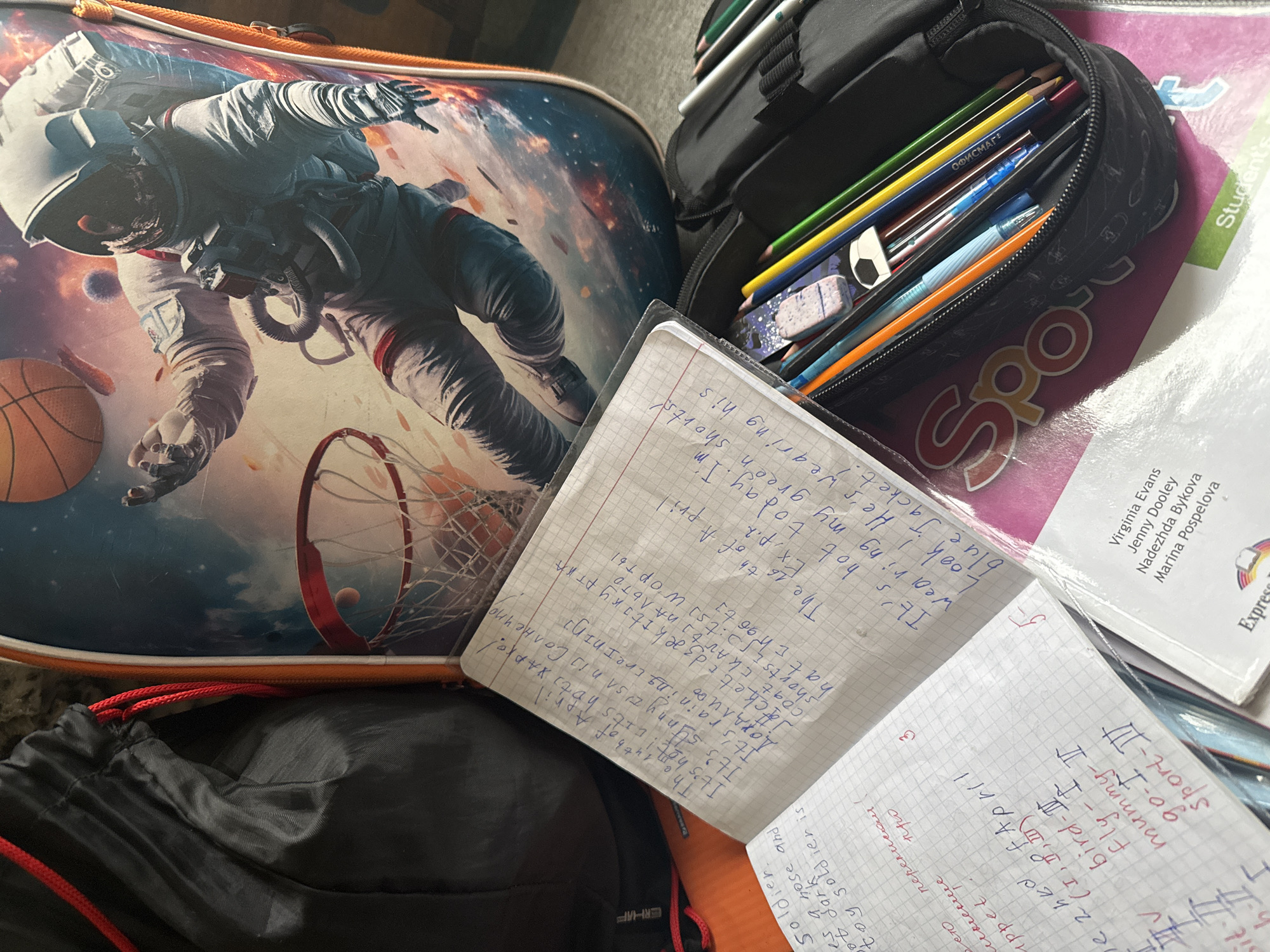 The image captures a close-up, top-down view of various school supplies, indicative of a student's workspace in Dmitrov, Russia.
The primary subject on the left is a vibrant school backpack or bag featuring a graphic of an astronaut in a space suit playing basketball in space, complete with a basketball and hoop. The bag has an orange trim. To its right, an open black pencil case is filled with an assortment of colored pencils, pens (including blue and orange ones), and an eraser shaped like a football.
Several notebooks and a textbook are scattered across the scene. An open, gridded notebook displays handwritten entries in blue ink, mixing English and Russian words, possibly a language learning exercise. Visible text includes phrases like "The 14th of April," "It's hot! It's hot," "It's sunny / Солнечно," "It's raining / Дождливо," and vocabulary such as "Jacket," "coat," "нальто" (coat), and "шорты" (shorts). Another entry reads, "The 16th of April, It's hot today. I'm wearing my green shorts. Look! He's wearing his blue Jacket!"
Beneath the pencil case, a textbook titled "Spot" is partially visible, listing authors Virginia Evans, Jenny Dooley, Nadezhda Bykova, and Marina Pospelova, suggesting it is an English language learning resource. Another gridded notebook, partially obscured and rotated, contains handwritten lists including "Sport-III," "go-I," "mummy-II," "bird-II," "Fly-I," and words like "puppet," "Soldier," "toy soldier is," and "переписом" (by re-writing/census). A black drawstring bag with red cords is visible at the bottom left.
The overall scene suggests a recently used or active study area, reflecting a student's daily academic life.
