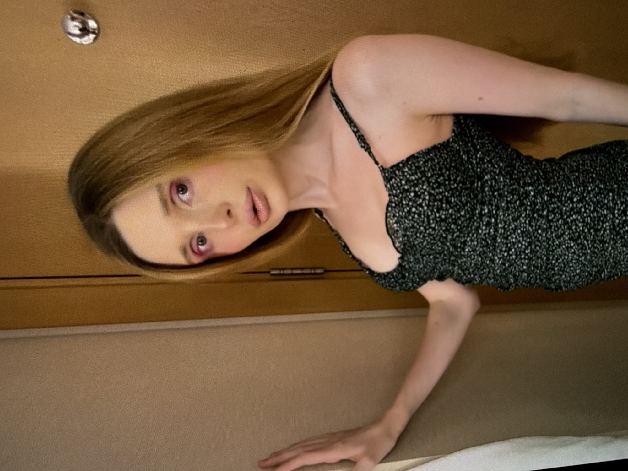 The media file shows a person with blonde hair and fair skin. They are wearing a black and white floral-patterned dress with thin straps. The person is looking towards the camera with a neutral expression. Their makeup includes pink eyeshadow and lipstick. The background appears to be indoors, with wooden paneling and a textured wall. There is a circular metallic object visible on the wooden paneling. The lighting suggests it is daytime, but no specific time can be determined. There is no visible text in the image.
