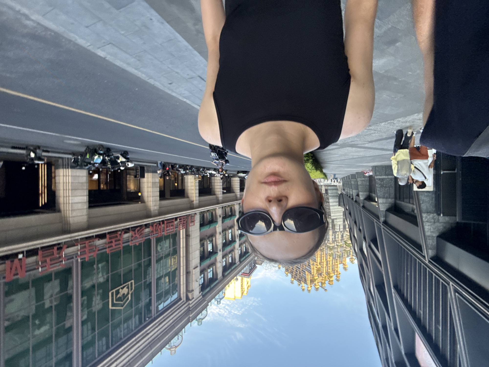 The image presents an inverted selfie taken by a person standing on a city street, looking down. The main subject, a person wearing a black tank top and sunglasses, occupies the foreground, with their head positioned at the bottom of the frame due to the upside-down orientation.
The background depicts a bustling urban street scene. On the left side of the frame (which would be the right side if the image were rotated correctly), a multi-story building with classical architectural features is prominent. Large red Chinese characters, clearly reading "中国银行" (Bank of China), are visible on its facade, alongside a distinctive corporate logo featuring a horse's head within a shield. Numerous motorcycles are parked along the sidewalk in front of this building.
On the opposite side of the street, a modern, dark-colored high-rise building with a sleek, angular design stretches upward. Further in the distance and reflected in some windows, a unique golden, ornate building with a complex, spired roof structure stands out against a clear blue sky dotted with scattered white clouds. Other pedestrians can be seen walking on the sidewalks on both sides of the street, some carrying bags. The scene is illuminated by bright daylight, suggesting a sunny day. The setting is an active urban environment, located in Putuo District, China.