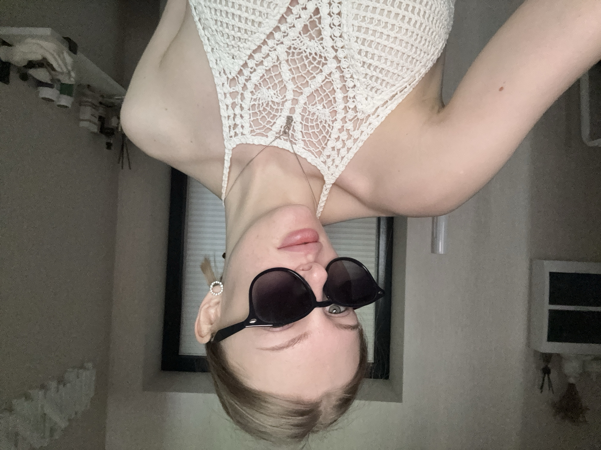A fair-skinned individual wearing a white crochet top and black sunglasses is photographed upside down in what appears to be an indoor setting. The person has light-colored hair. A silver necklace is visible. The background includes a window with closed blinds, a shelf with various items, and a white cabinet with hanging keys. The location cannot be determined as Perm, Russia based on the visual information provided.