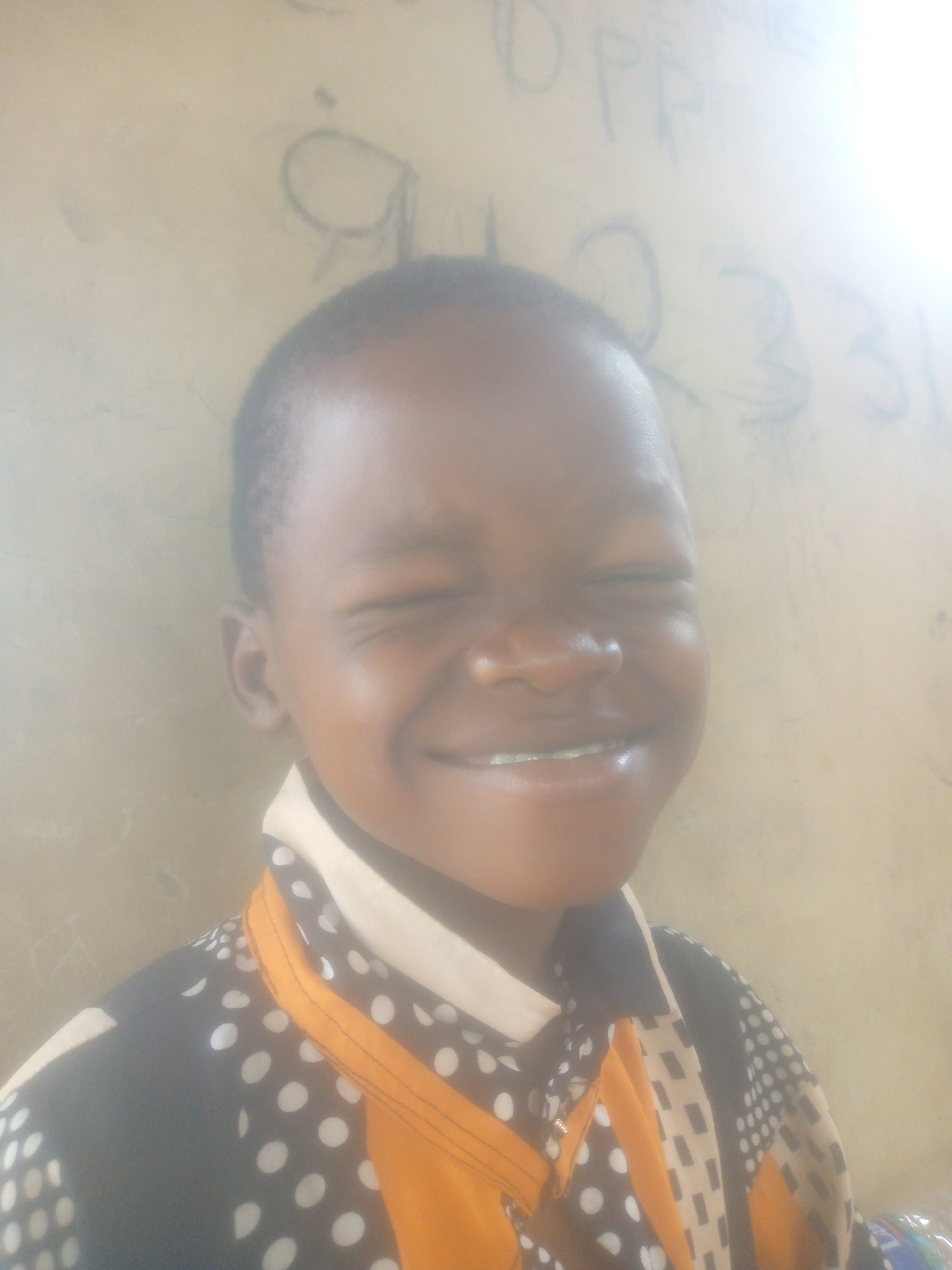 A close-up, brightly lit portrait captures a young child, likely a boy, smiling broadly with their eyes squinted shut, suggesting genuine joy or reacting to a bright light. The child has a dark skin tone and short, dark hair. They are wearing a collared shirt with a distinctive pattern featuring dark blue or black fabric with white polka dots, accented with orange stripes and lighter-colored sections containing dark rectangular dots. A small, metallic glint, possibly from dental braces or an object on a tooth, is visible on one of the upper front teeth.
The background consists of a light-colored, plain wall, possibly cream or light yellow. Faint markings, including handwritten letters and numbers, are visible on the wall. Partial letters like "D P P" or "D PR" can be seen near the top right, along with numbers that appear to be "2 3 3" or similar, perhaps "2331". A large, rounded shape resembling a "Q" or "9" is also discernible on the left side of the background. The lighting is very bright, casting a strong highlight on the right side of the image, which causes some overexposure and makes the background details hazy. The overall mood is one of happiness and cheerfulness.