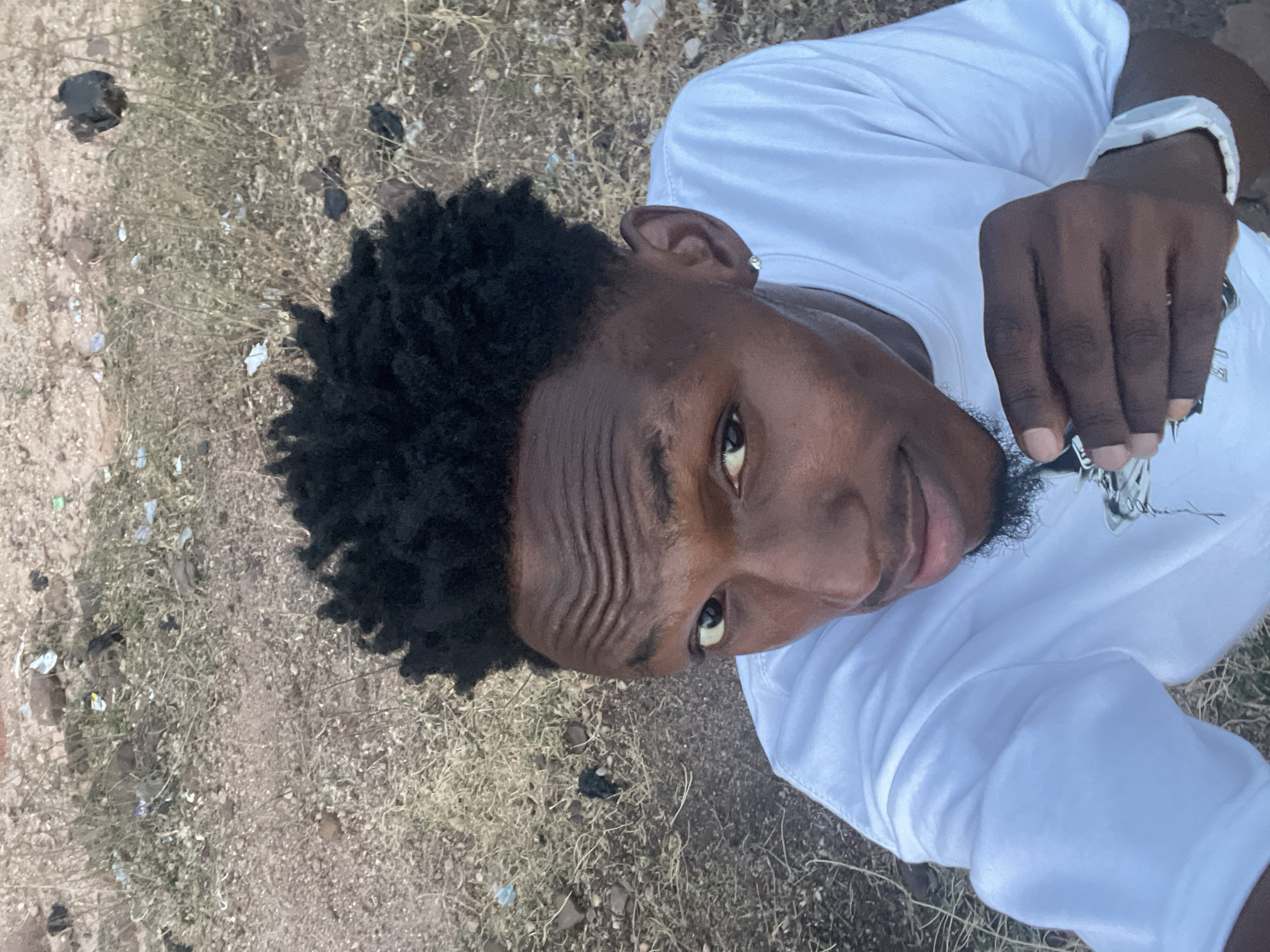 A medium-shot, high-angle portrait captures a young Black man looking directly up at the camera. He has short, dark, textured hair and a faint goatee. Prominent horizontal wrinkles are visible on his forehead. He wears a plain white t-shirt, a white digital watch on his left wrist, and a silver stud earring in his right ear. His right hand, positioned near his chest, appears to be holding a set of keys or a keychain.
The background is a dry, outdoor area with light brown, dusty soil mixed with small pebbles and sparse, dry grass and weeds. Several pieces of light-colored trash, such as plastic fragments or paper, are scattered across the ground, indicating an unkempt environment. The lighting suggests it is daytime, with clear visibility. The man's expression is neutral, focusing intently on the camera. The scene takes place in Lokoja, Nigeria. There is no discernible text visible in the image.